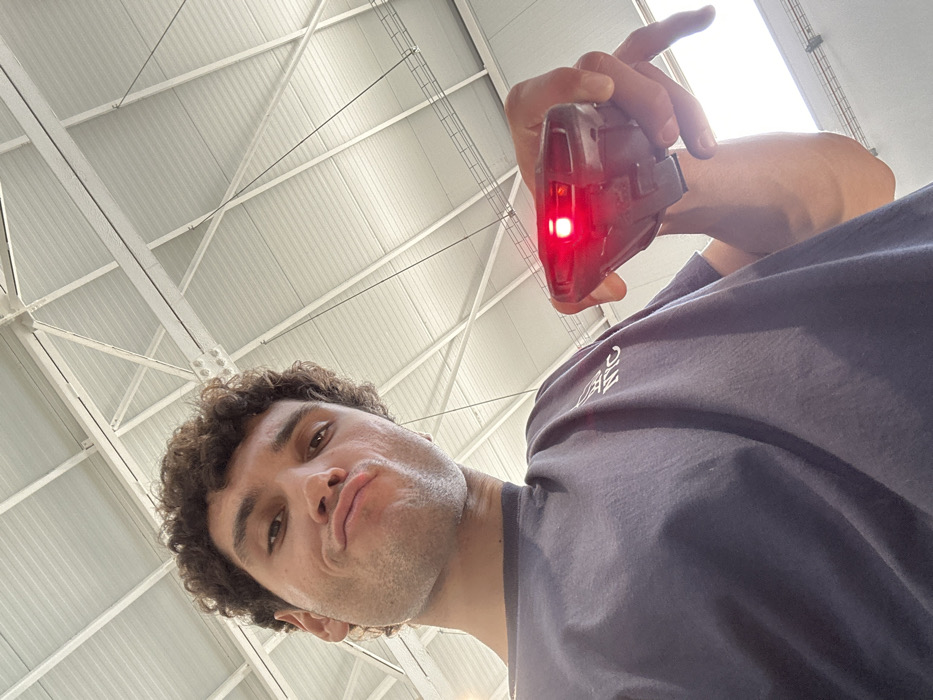 The image depicts an adult male taking a selfie within an industrial setting in Polígon Industrial De Son Castello, Spain. The male, visible from the chest up, has curly brown hair and light skin, and is making a pouting facial expression directly towards the camera. He is wearing a dark blue t-shirt with white lettering on the left chest. His right hand holds a black, angular electronic device with a prominent, illuminated red light on its front face, positioned in the lower right portion of the frame. The background features a high, light-colored ceiling composed of a metal truss structure and corrugated panels, characteristic of a warehouse or large industrial building.