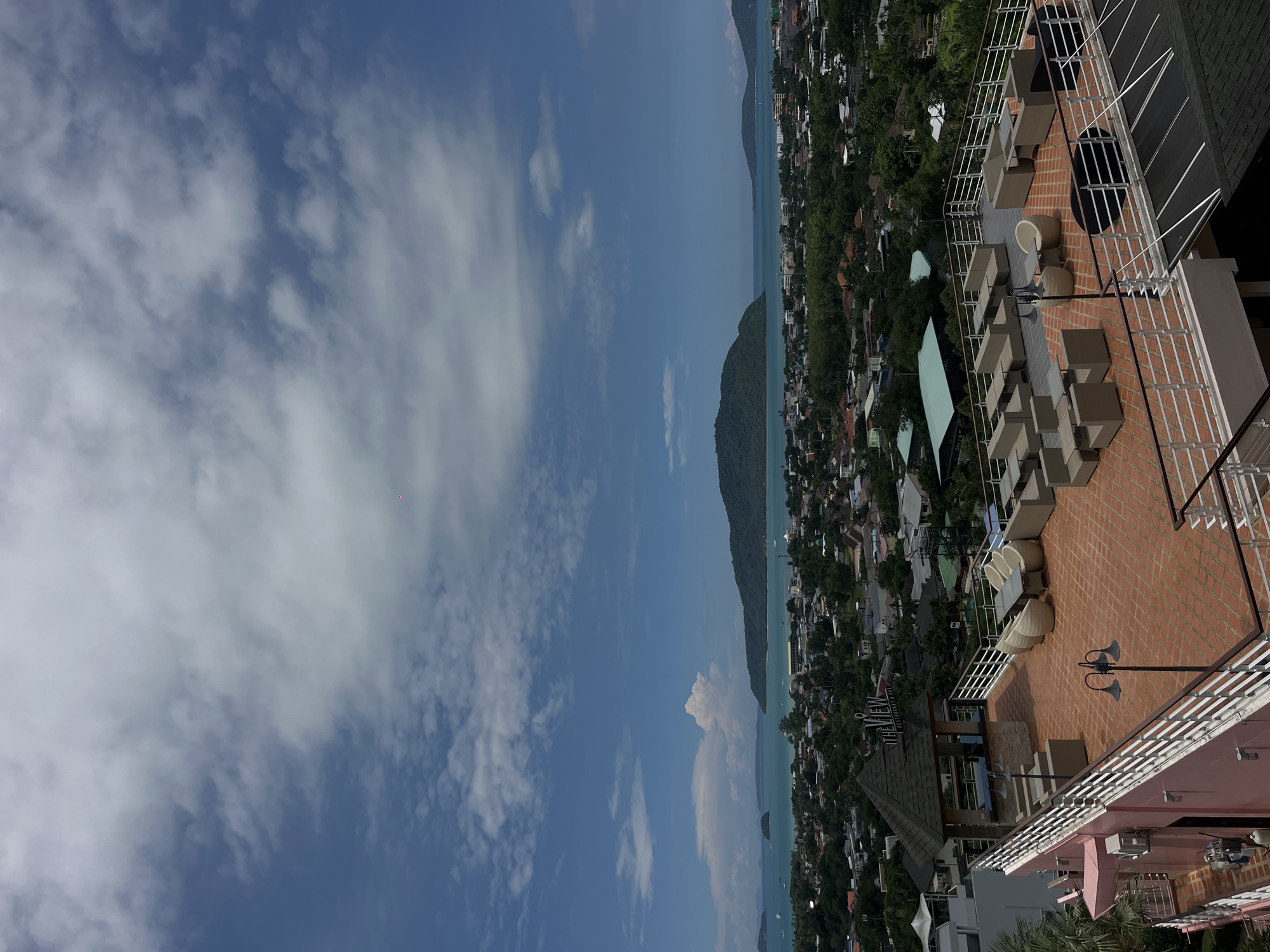 This is a high-angle, eye-level shot of a rooftop restaurant and bar area overlooking Rawai, Thailand. The rooftop is furnished with outdoor seating, including modular sofas, armchairs, and tables, arranged for dining and lounging. The flooring of the rooftop is tiled with terracotta-colored tiles. A covered gazebo-like structure with the sign "THE VIEW RESTAURANT BAR" is visible on the rooftop. The background features a panoramic view of Rawai, with a dense cityscape of buildings and lush green trees, extending to the turquoise sea. Several islands and a larger, mountainous island are visible in the distance, under a partly cloudy blue sky. No people are visible in the image.