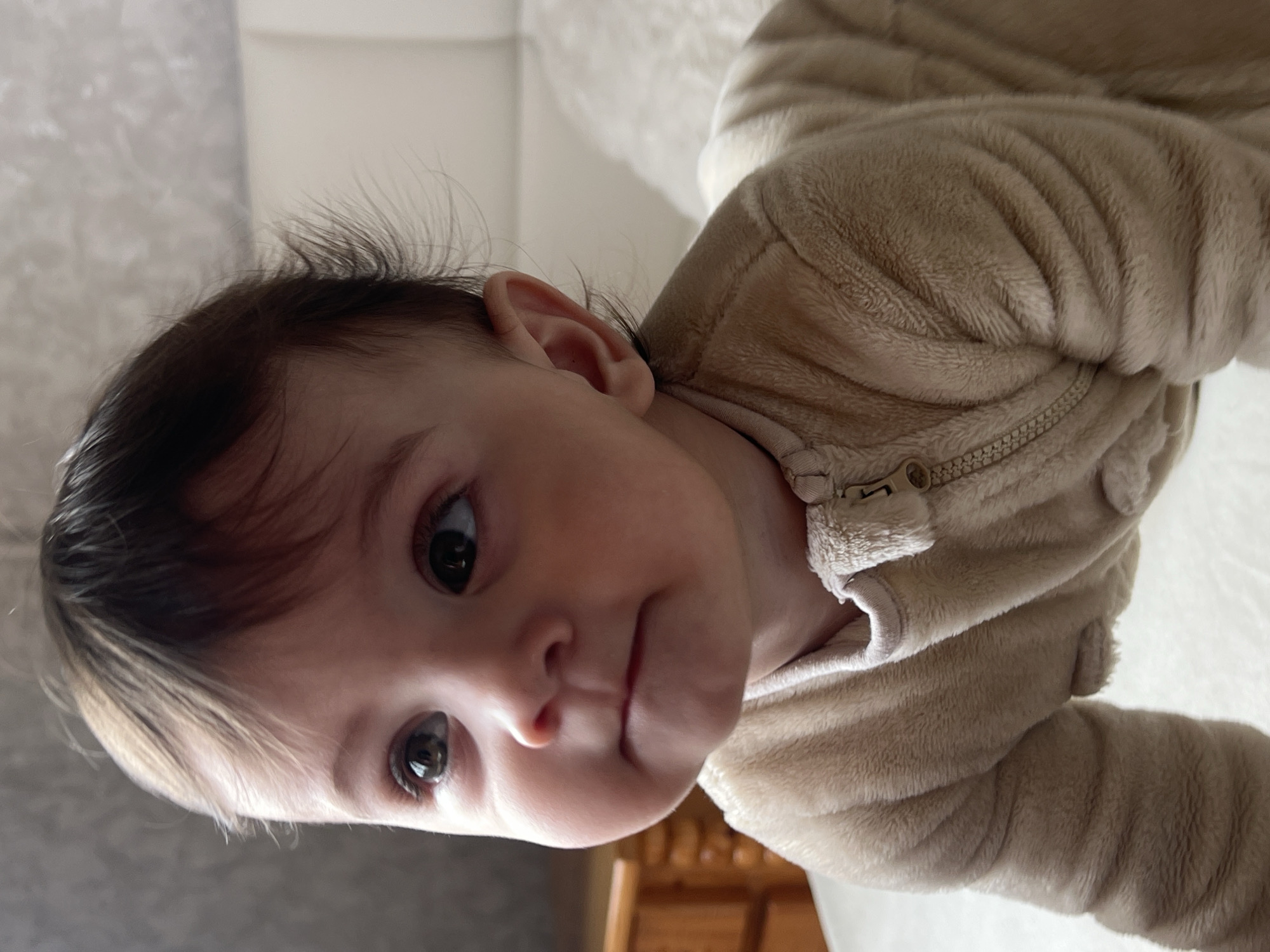 This image captures a close-up of a single infant in a domestic setting within the city of Elena, Bulgaria. The subject is a baby with fair skin, dark brown hair, and large dark eyes, looking upwards and to the left of the frame with a neutral expression and slightly parted lips. The baby is dressed in a light beige, plush, long-sleeved one-piece garment, likely a sleeper, featuring a visible zipper pull on the upper chest. The posture suggests the infant is in a crawling or pre-crawling position, with the upper body slightly elevated. In the blurred background to the left, a light-colored wooden furniture piece, possibly part of a crib or dresser, is partially visible. To the right and behind the infant, a light-colored, possibly textured wall or headboard is present, alongside a soft, light-colored fabric surface, indicating a bed or padded area. The lighting is soft and ambient, illuminating the infant's face from the front-right.