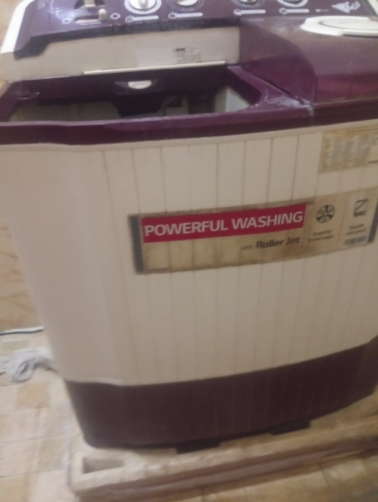 The image shows a top-loading washing machine. The machine is two-toned, with a light cream body and a burgundy top and base. A label on the side reads "POWERFUL WASHING with Roller Jet". The washing machine appears to be located indoors on a tiled floor. No people are visible. The location is Aviation Quarters, Nigeria.