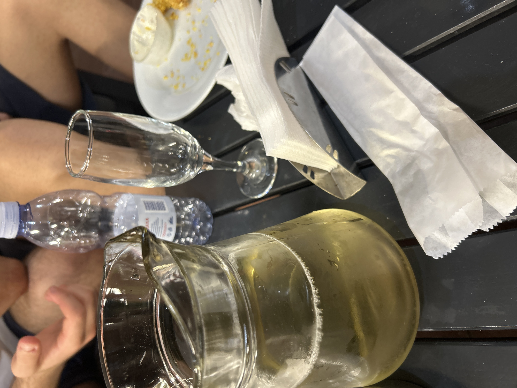 The image depicts a dining table scene in Blagoevgrad, Bulgaria, featuring a dark, slatted table surface. In the foreground, a white paper packet lies to the right of a silver-toned metal napkin dispenser filled with white paper napkins. To the left, a clear glass pitcher contains a pale yellow liquid with a visible layer of bubbles at the surface. Adjacent to the pitcher, an empty, clear stemmed glass stands upright. Behind the pitcher, a partially full clear plastic bottle with a blue cap is visible, marked with "500ml" on its label. On the right side of the table, a white ceramic plate holds a small white ramekin containing a creamy white substance, along with scattered yellow food crumbs and a small portion of a golden-brown fried item. In the background, the lower arms and hands of one individual are visible on the left, while the legs of another person are partially seen on the right, wearing dark apparel.