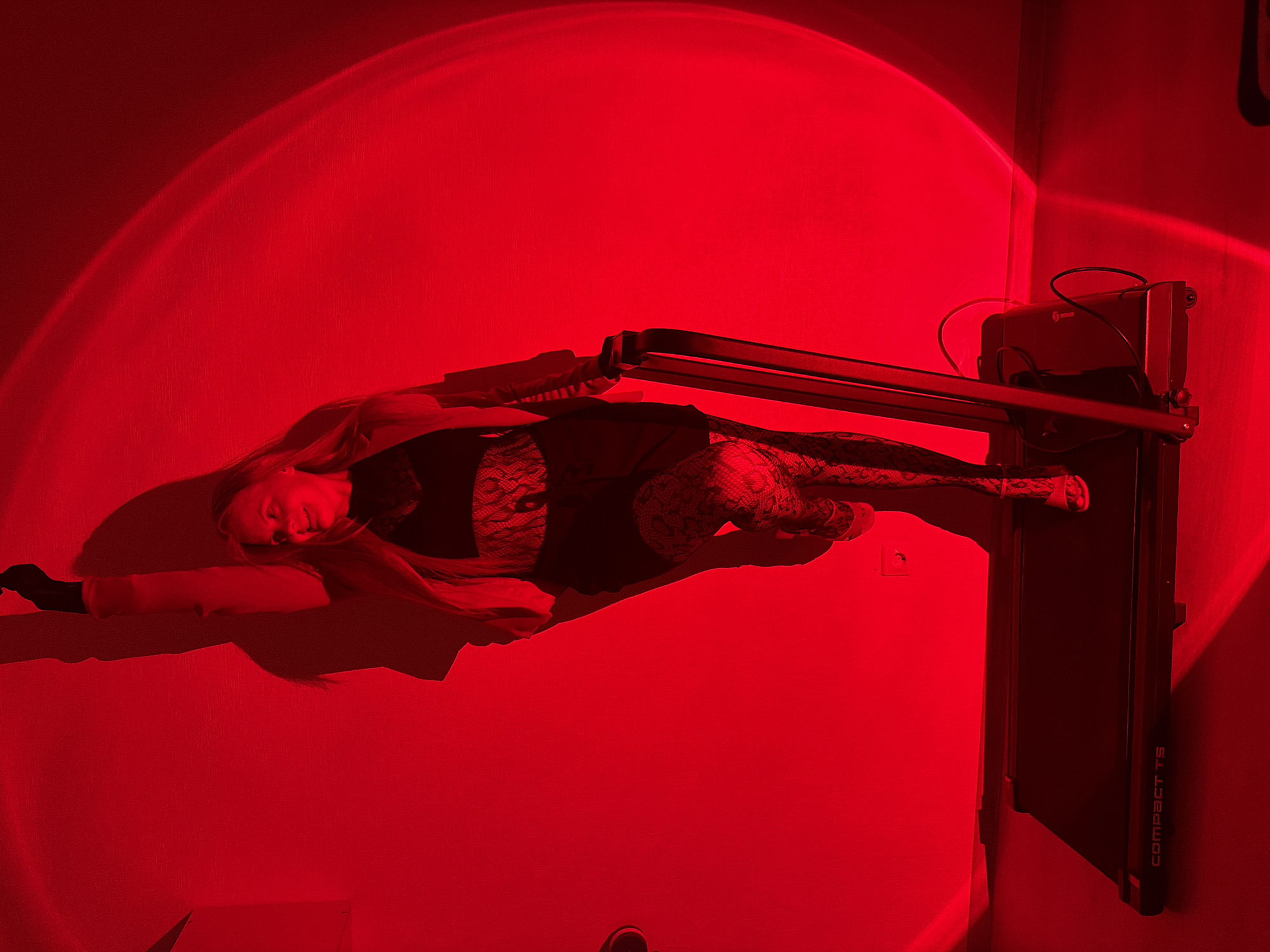 In a room illuminated by a strong red light, a woman with blonde hair stands on a black treadmill. She is wearing a black lace top, a black skirt, and patterned tights. Her left arm is raised, and her right hand is on the treadmill's handlebar. The treadmill displays the text "COMPACT T5". The red light casts a large circular glow on the wall behind her, and a shadow of her is visible on the same wall. The environment appears to be indoors, with a wooden floor visible beneath the treadmill. There are no discernible details suggesting a specific location within the city of Izhevsk, Russia.