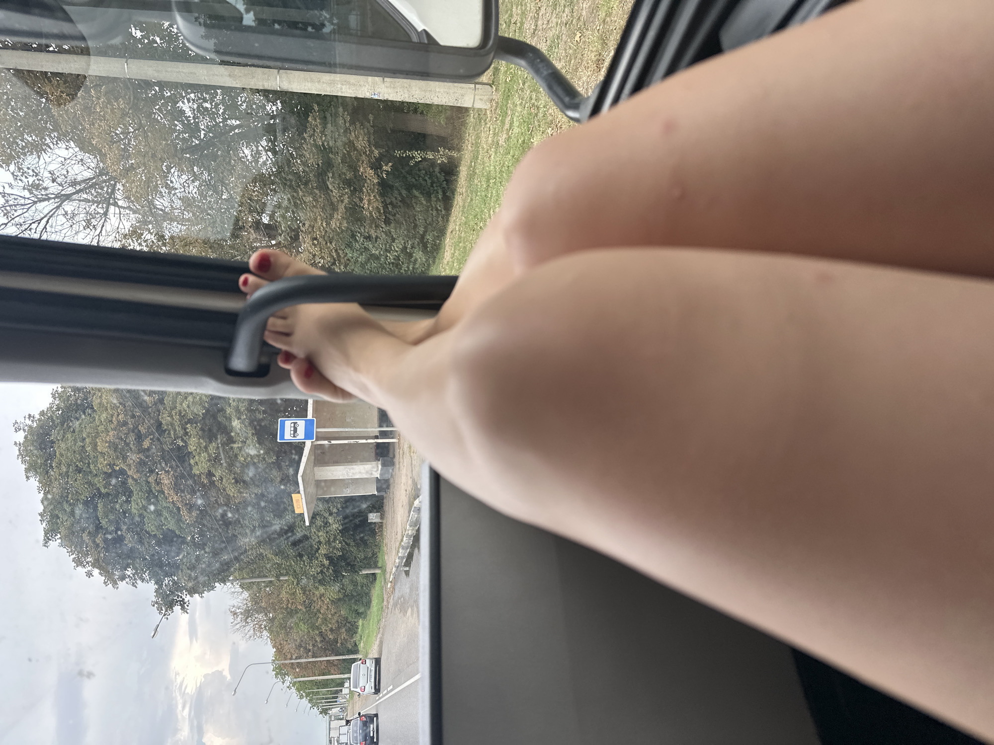 The image shows a view from inside a vehicle, looking out the window. In the foreground, a person's bare legs and feet are visible, with their legs crossed. The feet are propped up on something within the vehicle. Outside the window, a road is visible with cars driving on it, and a bus stop with a shelter and a sign is present. Trees and vegetation line the road. The sky is cloudy. The scene appears to be along a roadside in the city of Novotitarovskaya, Russia, given the presence of a bus stop and typical roadside scenery.