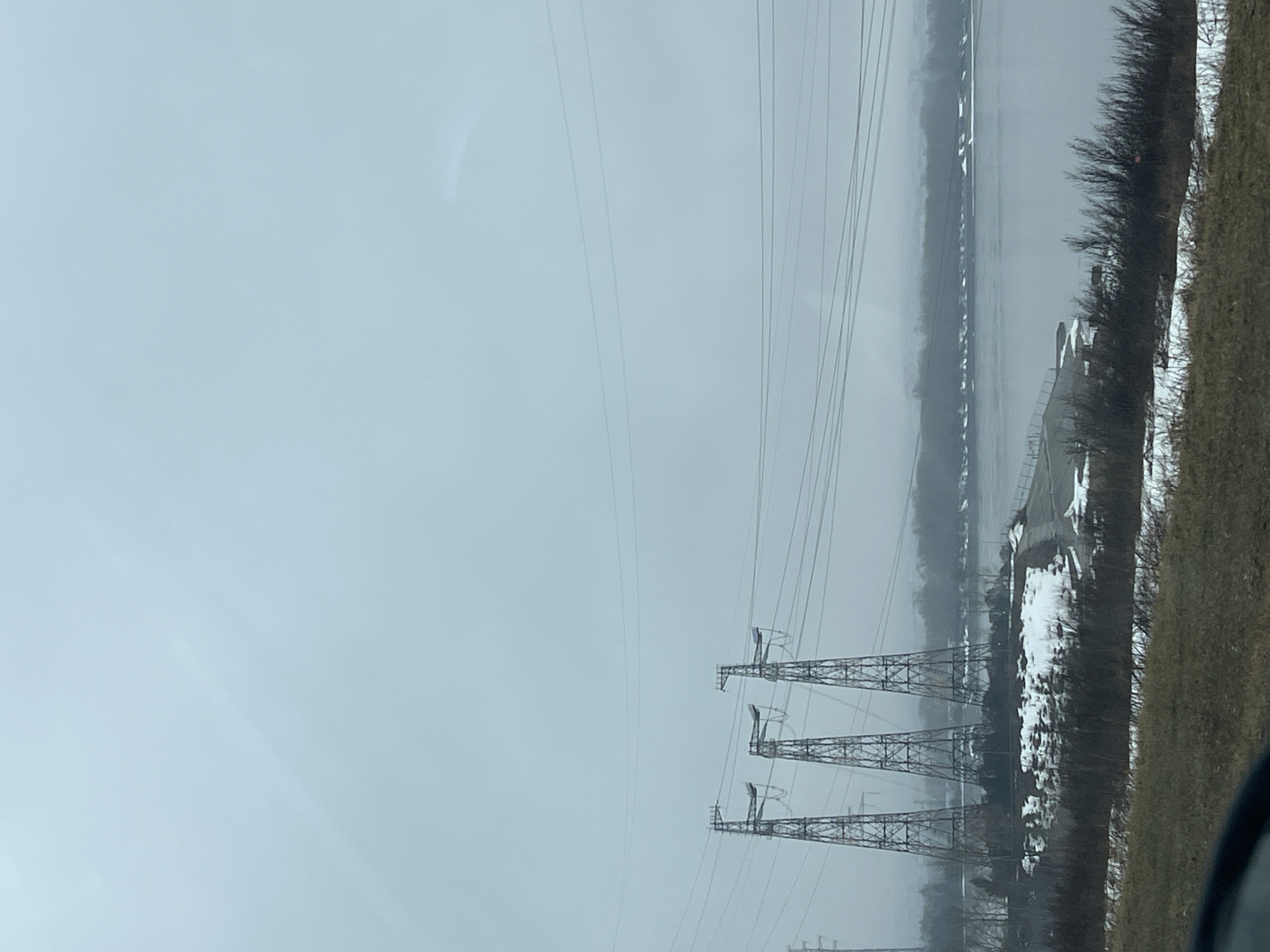 The image depicts an overcast landscape in what is likely Chaikovsky, Russia, given the presence of the Kama River. Three large metal transmission towers are in the foreground, carrying numerous power lines. To the right, the edge of the Kama River can be seen, with visible snow along the riverbank and vegetation. The background is obscured by fog or haze, further diminishing visibility. The foreground appears to be a field or embankment, as seen by some vegetation and the view from a car window in the lower right corner. The scene suggests a cold, winter or early spring season.