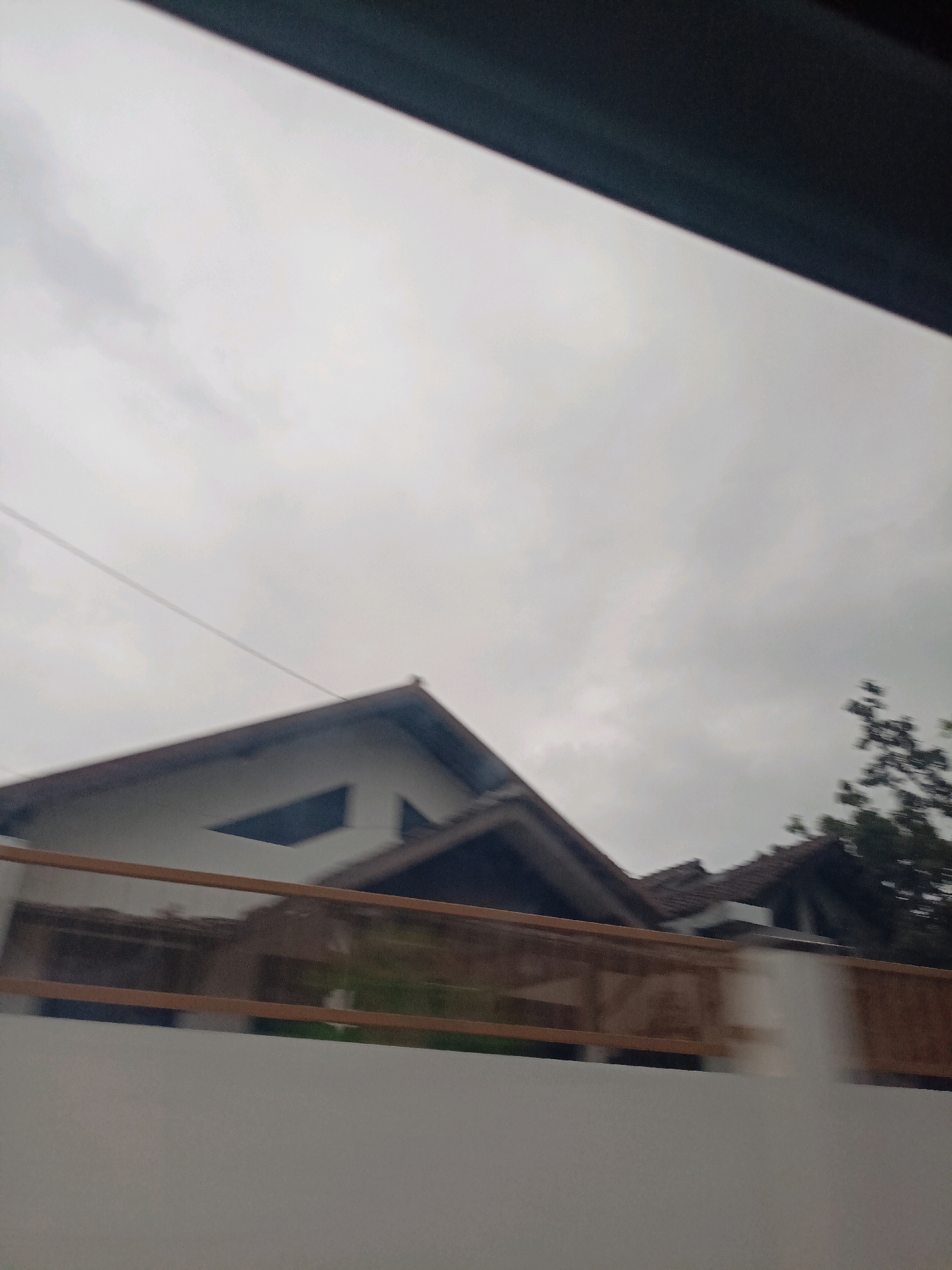 The image captures a cloudy sky over a residential area in Magelang, Indonesia. In the foreground, a light-colored wall runs horizontally, with a wooden fence above it. Beyond the fence, two houses are visible, characterized by their sloped, tiled roofs and light-colored facades. One house features a distinct triangular dark window. To the right, a tree with dark green foliage can be seen. A thin, dark wire stretches diagonally across the sky. The overall impression is of an overcast day in a suburban setting.