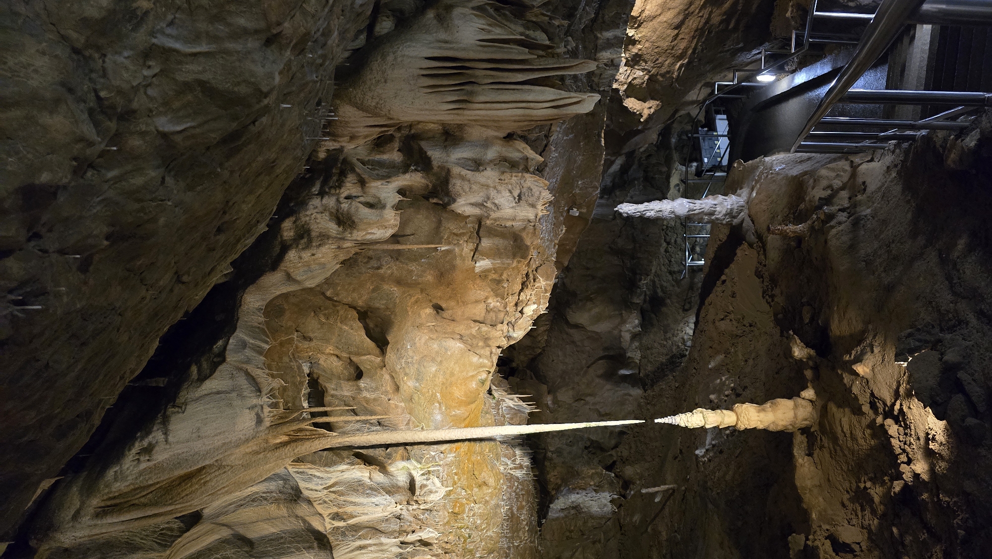 This image depicts the interior of a cave, likely a tourist attraction given the presence of a metal railing and stairs in the background. The cave walls are rough and textured, with formations of stalactites and stalagmites visible throughout. In the foreground, a prominent stalactite hangs horizontally, illuminated by a light source that casts a distinct glow. Several smaller stalactites are also visible, appearing as white, needle-like structures on the cave walls and ceiling. The lighting is artificial, designed to highlight the cave's natural features. The overall scene conveys a sense of awe and natural wonder. The location is identified as Vavřinec, Czechia, which likely refers to a specific cave system in that region. There is no visible text in the image.