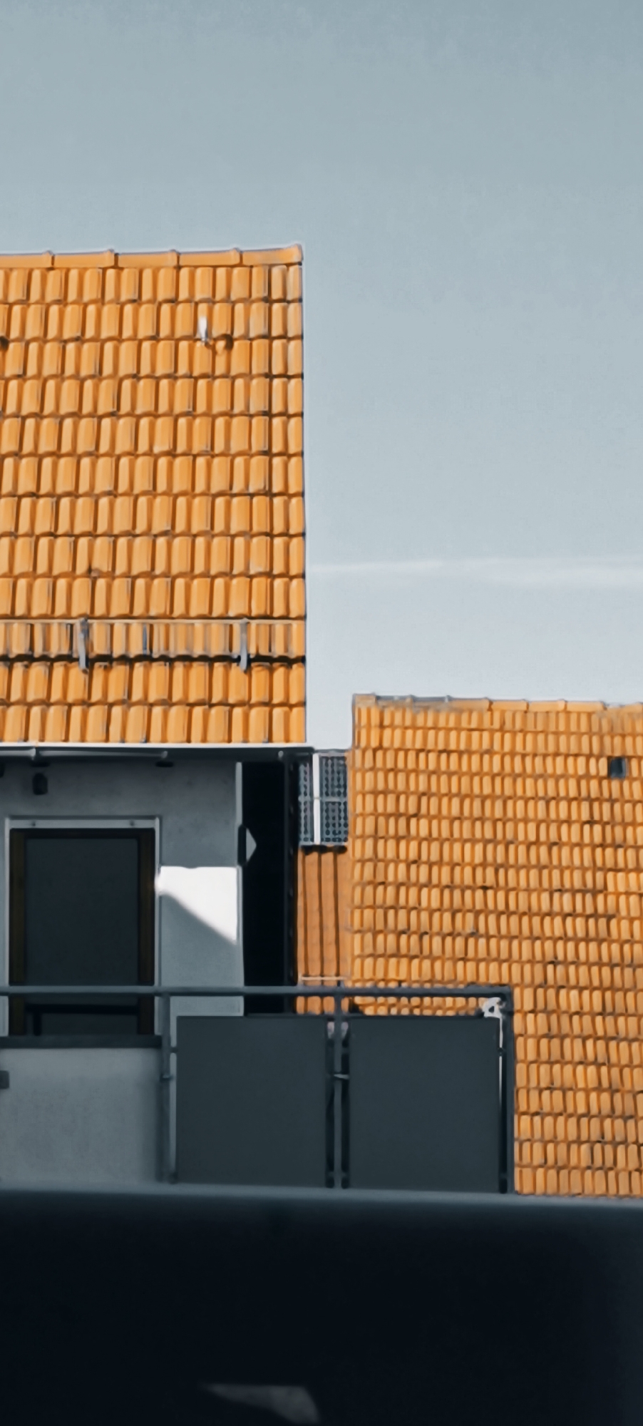The image shows a close-up view of two adjoining buildings with distinctive orange tile roofs, set against a pale blue sky. The building on the left features a white facade with a dark-framed window and a section of a balcony with dark grey railings. The roofline of this building is clearly visible, composed of rows of overlapping orange tiles. To the right, a portion of a second building is visible, also capped with a similar orange tile roof. This second building appears to be constructed of lighter colored blocks and has a smaller window with a grate. The overall impression is of a residential area, possibly in a town or city, given the architectural style. The lighting suggests it is daytime, with shadows cast on the white wall of the left building. This image was taken in Winnenden, Germany.