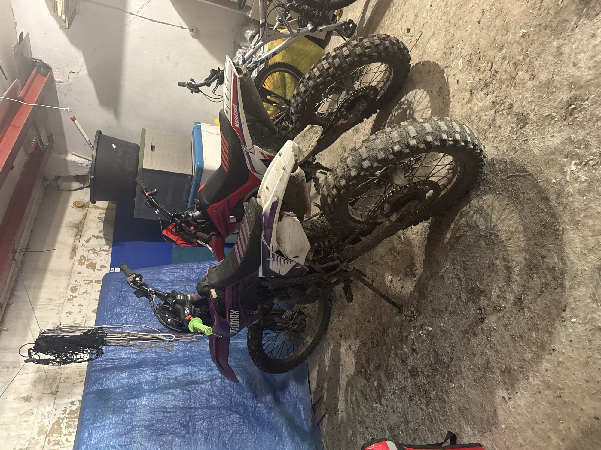 This media file shows two dirt bikes parked indoors. The bikes are oriented in a way that makes it difficult to determine the exact number of motorcycles, but at least two are visible. The scene appears to be a garage or storage area, with a concrete floor covered in dirt and debris, and white walls. One of the bikes is predominantly red and white, with the branding "Motoland" visible on its side. The other bike is purple and white. Both motorcycles have knobby tires, suggesting they are off-road vehicles.
In the background, there are various objects including a large black bucket, a blue tarp, and parts of other equipment that are not clearly identifiable. A bicycle is also partially visible behind the red and white dirt bike. The lighting is dim, indicating it might be nighttime or the area is poorly lit. There are no people present in the image, and the overall impression is one of a storage space for recreational vehicles. The location context of Novokuybyshevsk, Russia, does not offer any specific visual cues within the image itself. The image is characterized by a sense of clutter and a utilitarian setting, rather than any particular activity or event.
