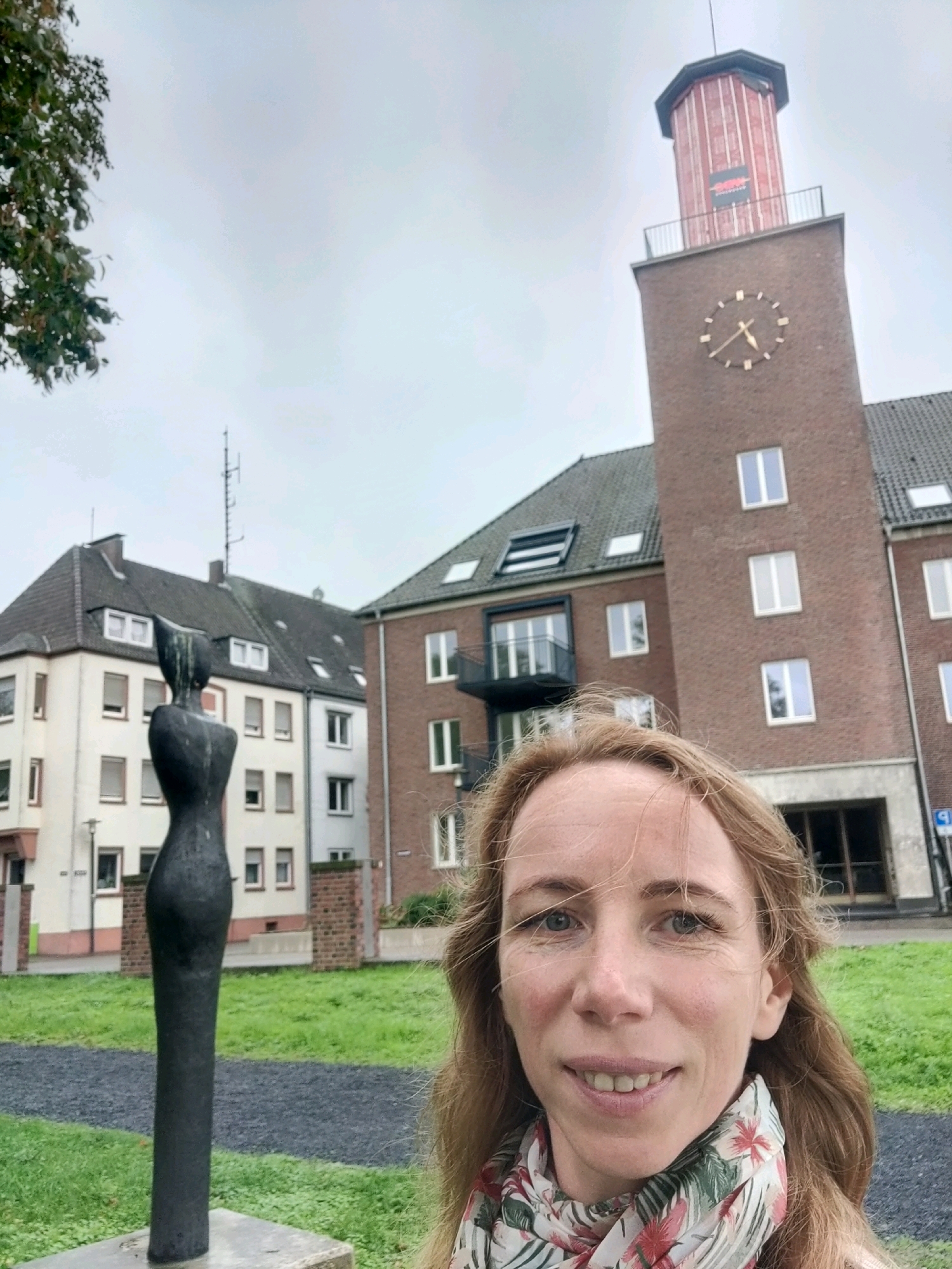 A woman with long, light brown hair and blue eyes smiles at the camera. She is wearing a floral scarf. Behind her, to the left, is a tall, dark, abstract sculpture. Further back, there are several buildings, including a brick tower with a clock. The sky is overcast. The scene appears to be an outdoor public space in the city of Emmerich on the Rhine, Germany.