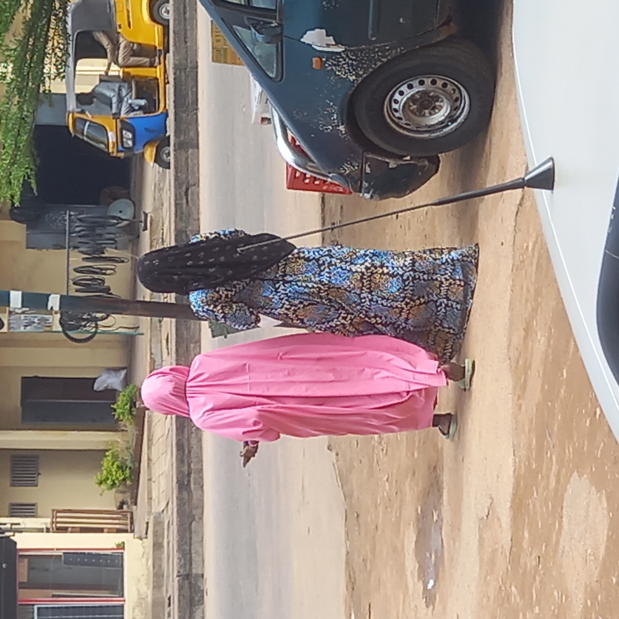 In Dadi Riba, Nigeria, two women are walking on a street next to several cars and a tricycle. One woman wears a pink hijab and robe and the other woman wears a floral dress with a dark-colored hair covering. The street is paved but worn with some discoloration. Buildings are visible in the background, including a light-colored building with horizontal bars and windows. Other cars are visible in the background.