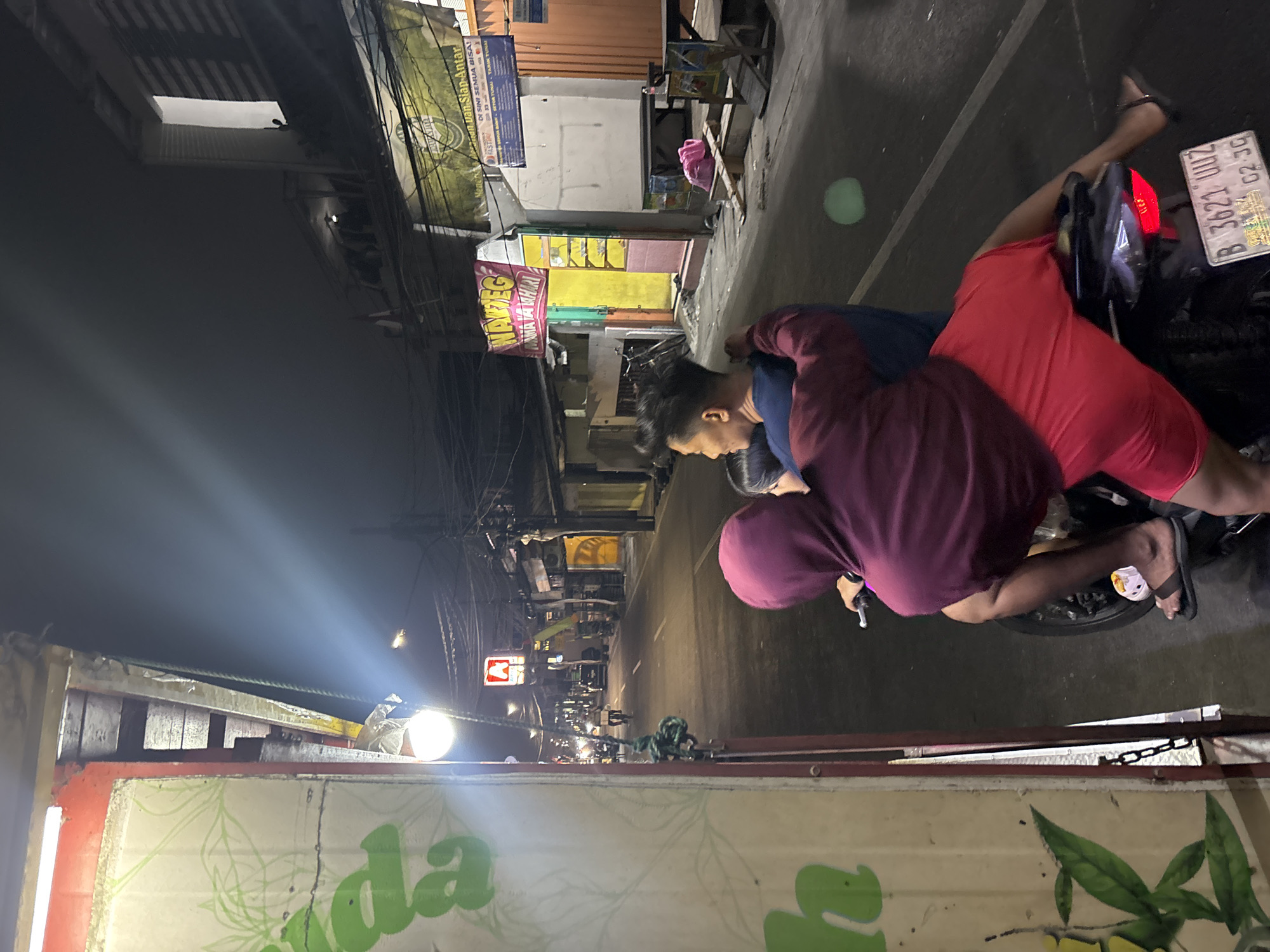 The image captures a nighttime scene on a street in North Jakarta, Indonesia, specifically in the Taman Sari area based on visual cues. In the foreground, two individuals are seated on a motorcycle. The person in the back, wearing a burgundy hoodie, has their arms around the person in front. The person in front is wearing a dark blue shirt and is looking to the left. The motorcycle has a license plate that reads "B 3621 UU2". The environment is an urban street with shops and businesses lining both sides. A brightly lit sign for "WARTEG" is visible on the left. Overhead, power lines and streetlights illuminate the scene, casting shadows and highlighting the nighttime atmosphere. The road appears to be paved and wet, possibly from recent rain. Several other vehicles and pedestrians are visible in the background, suggesting moderate street activity.