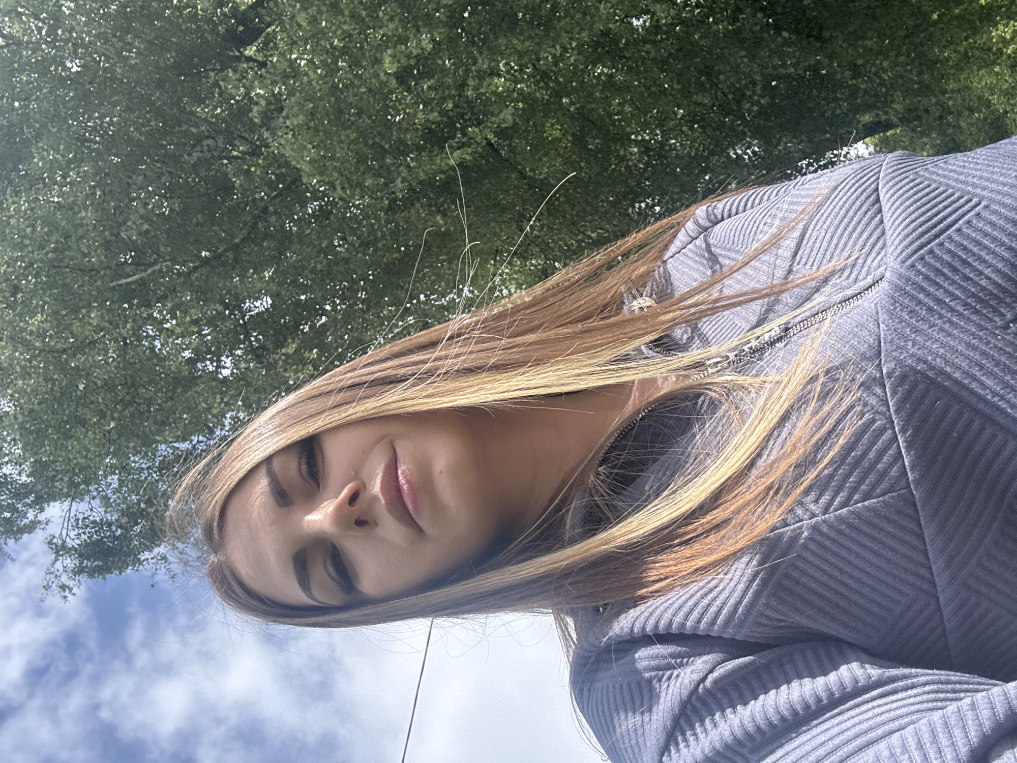 A young woman is pictured from a low angle, looking down at the camera. She has blonde hair with brown roots, and fair skin. She's wearing a gray ribbed jacket. Above and behind her is a canopy of green leaves, and glimpses of blue sky with white clouds are visible. The location in the city of Shevlyakovo, Russia, is unknown.
