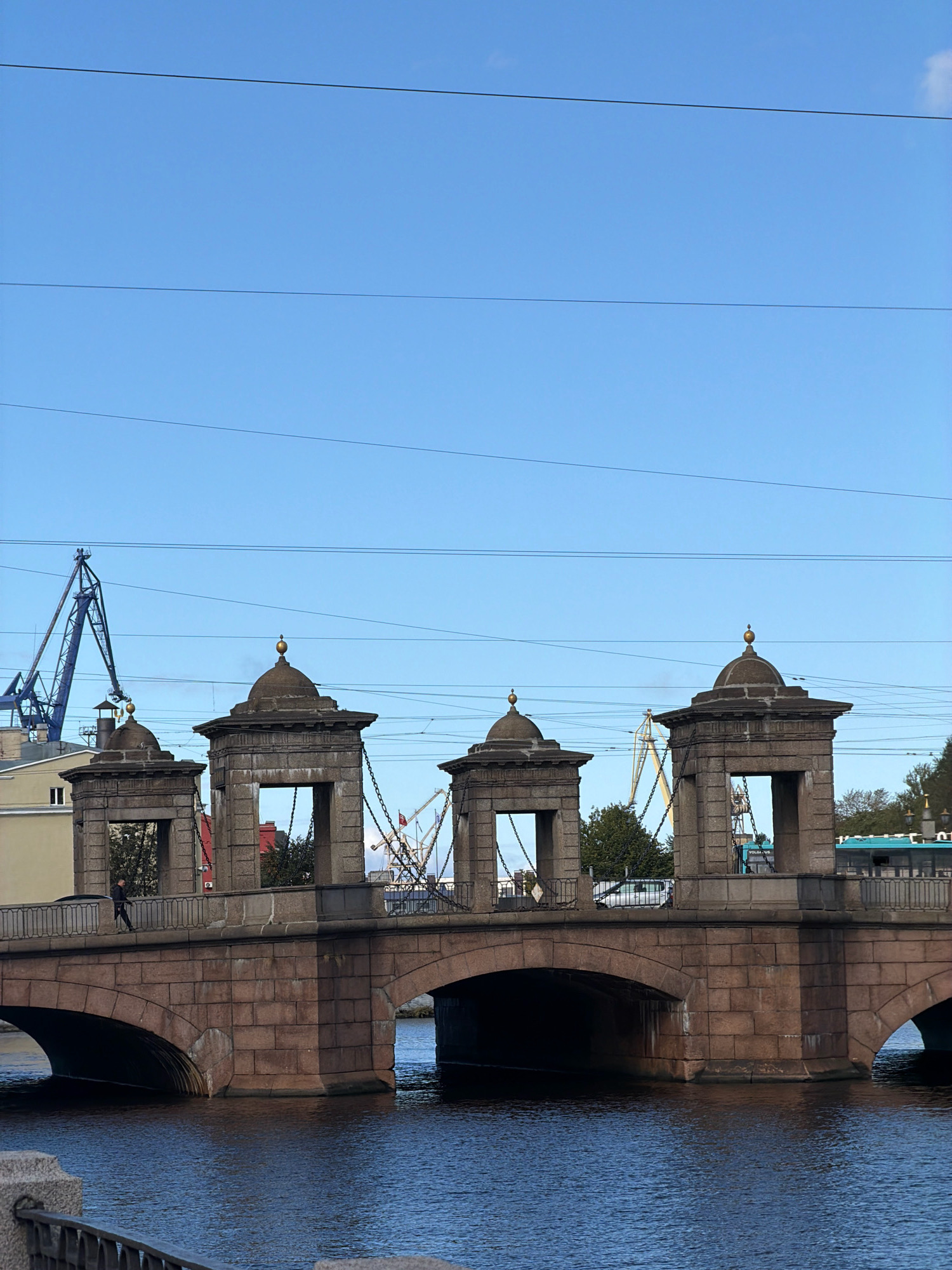 The image shows a view of the Anichkov Bridge in Saint Petersburg, Russia, spanning the Fontanka River. The bridge is characterized by its stone arches and the decorative pavilions at each corner, topped with domes. In the foreground, the dark blue water of the river reflects the overcast sky. On the bridge, a single pedestrian is walking. In the background, industrial cranes are visible against the clear blue sky, suggesting the presence of port facilities or construction nearby. The overall scene depicts a blend of historical architecture and industrial elements within the urban landscape of Saint Petersburg.