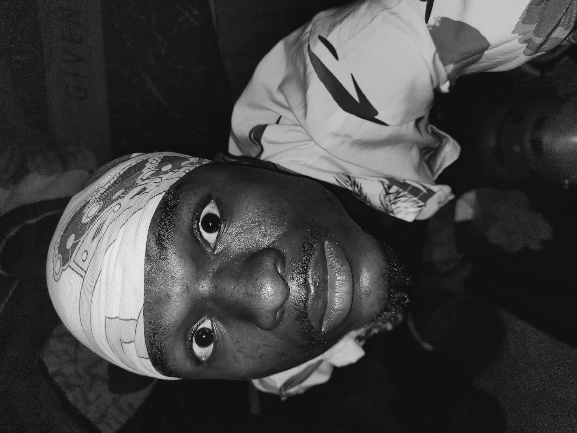 A black and white close-up, low-angle shot captures a young Black male wearing a patterned bandana and a patterned shirt. The man's eyes are wide open, and he appears to be looking directly at the camera. His face is lit from above, highlighting sweat on his forehead and cheeks. A faint mustache and beard are visible. The background is dark, with a blurred sign that appears to read "GIVEN." The image was taken in Bukuru, Nigeria, in what looks like an indoor setting with dim lighting.