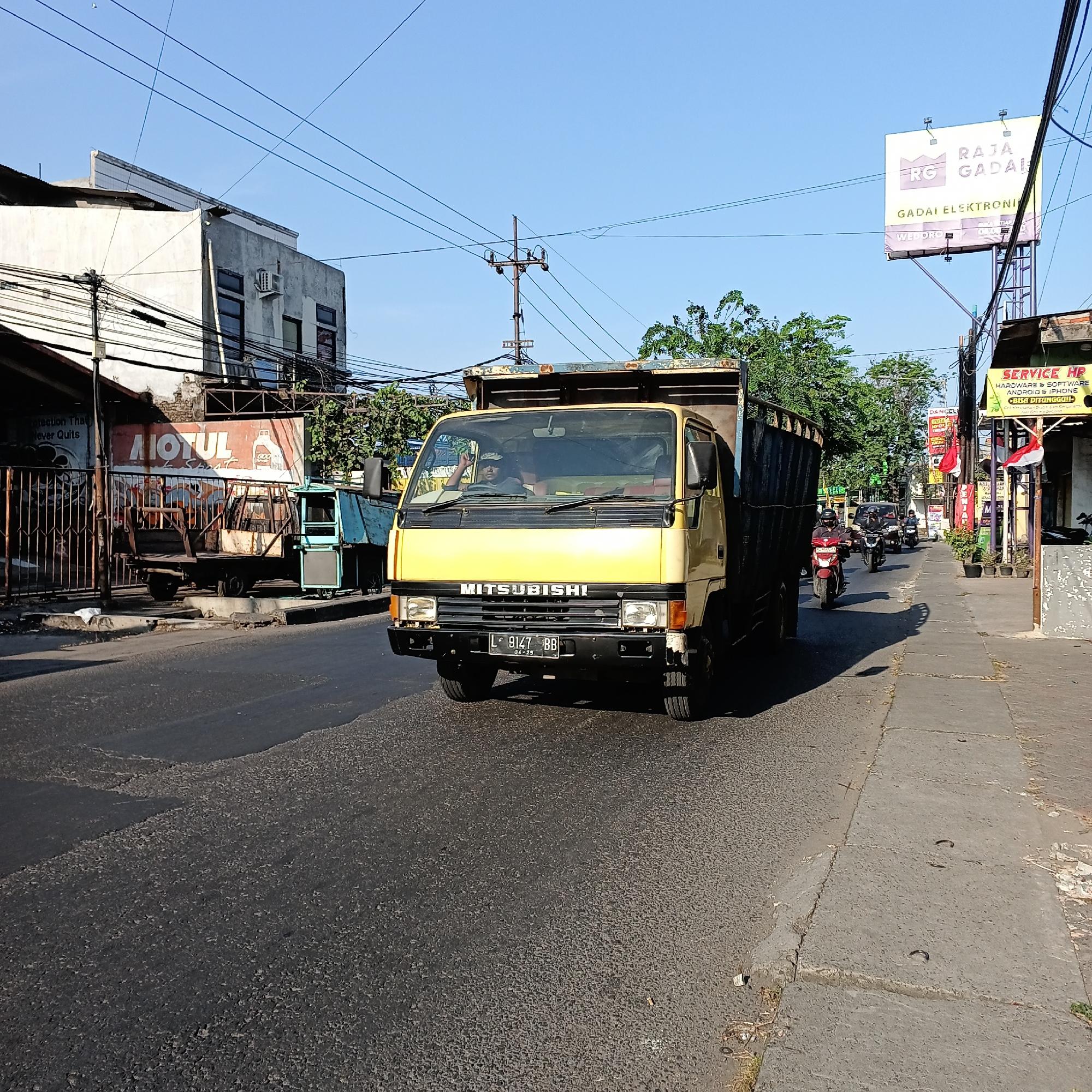 In Wedoro, Indonesia, a yellow Mitsubishi dump truck travels down a paved road under a clear blue sky. The truck's license plate, L 9147 BB, is visible. A person wearing a black cap is driving the truck. To the left of the road, a building with white and gray walls is visible, along with a metal fence and another parked vehicle. To the right, a sidewalk runs alongside the road, with a billboard advertising "Raja Gadai Elektronik" and a "Service HP" sign. Other vehicles, including motorcycles, are also on the road. Trees and electrical wires are present in the background.