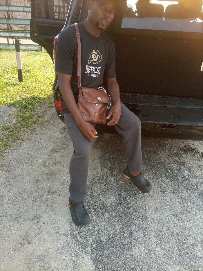 A dark-complexioned adult male sits on the open tailgate of a dark-colored SUV or van. He wears a black baseball cap, a black t-shirt featuring a "BUFFALOES COLORADO" logo, grey pants, and black clogs with orange accents. A brown leather messenger bag hangs over his right shoulder, resting on his lap, with white earphones visible near it. He has a beard and looks directly towards the camera with his mouth slightly open. The ground is a light-colored, gravelly surface. To the left, green grass and a black and white striped pole are present. The specific location within Eliozu, Nigeria, is not discernible from the image details.