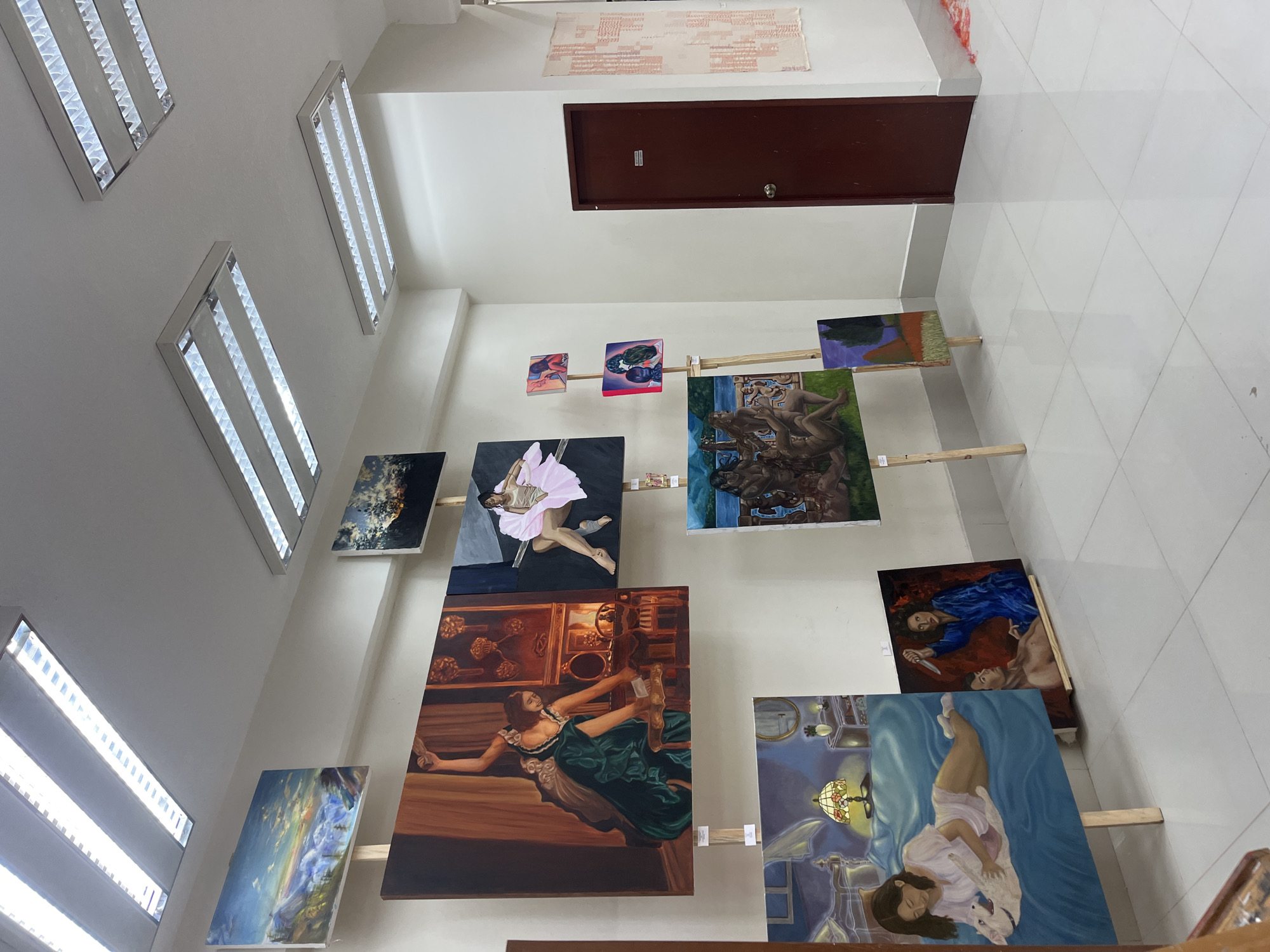 The image captures an interior space in what appears to be a gallery or exhibition area. The location is San Andrés Cholula, Mexico. The room is brightly lit by multiple rectangular fluorescent lights along the wall near the ceiling. The walls are painted white, and the floor is covered with large, light-colored tiles.
Several paintings are displayed along the walls using wooden supports. The paintings vary in size and subject matter, including figurative works, landscapes, and abstract compositions. Some paintings depict human figures in various poses and settings. A brown door is visible on one wall.
Above the door, a rectangular piece of patterned cloth or textile is mounted on the wall.