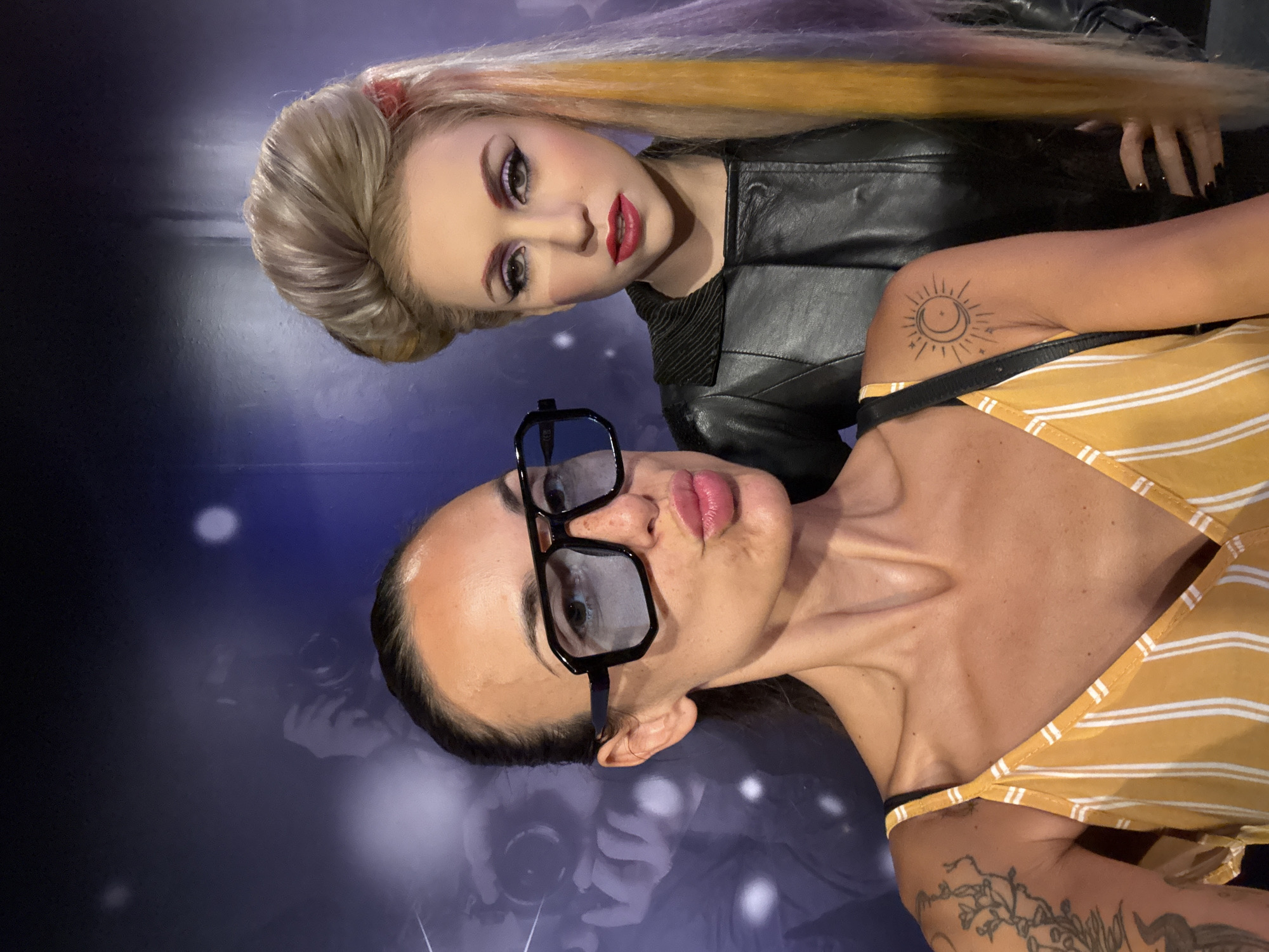 A woman with dark hair pulled back in a ponytail, wearing large black-rimmed glasses and a yellow and white striped top, poses for a selfie with a wax figure. The wax figure, positioned behind and to the right of the woman, is a blonde woman with dramatic makeup, including bold eyeliner and red lipstick, wearing a black leather jacket. The background appears to be a dark wall with a large, blurry image of photographers with cameras, suggesting a museum or a themed exhibit. The overall setting is indoors. The image was taken in Prague, Czechia.