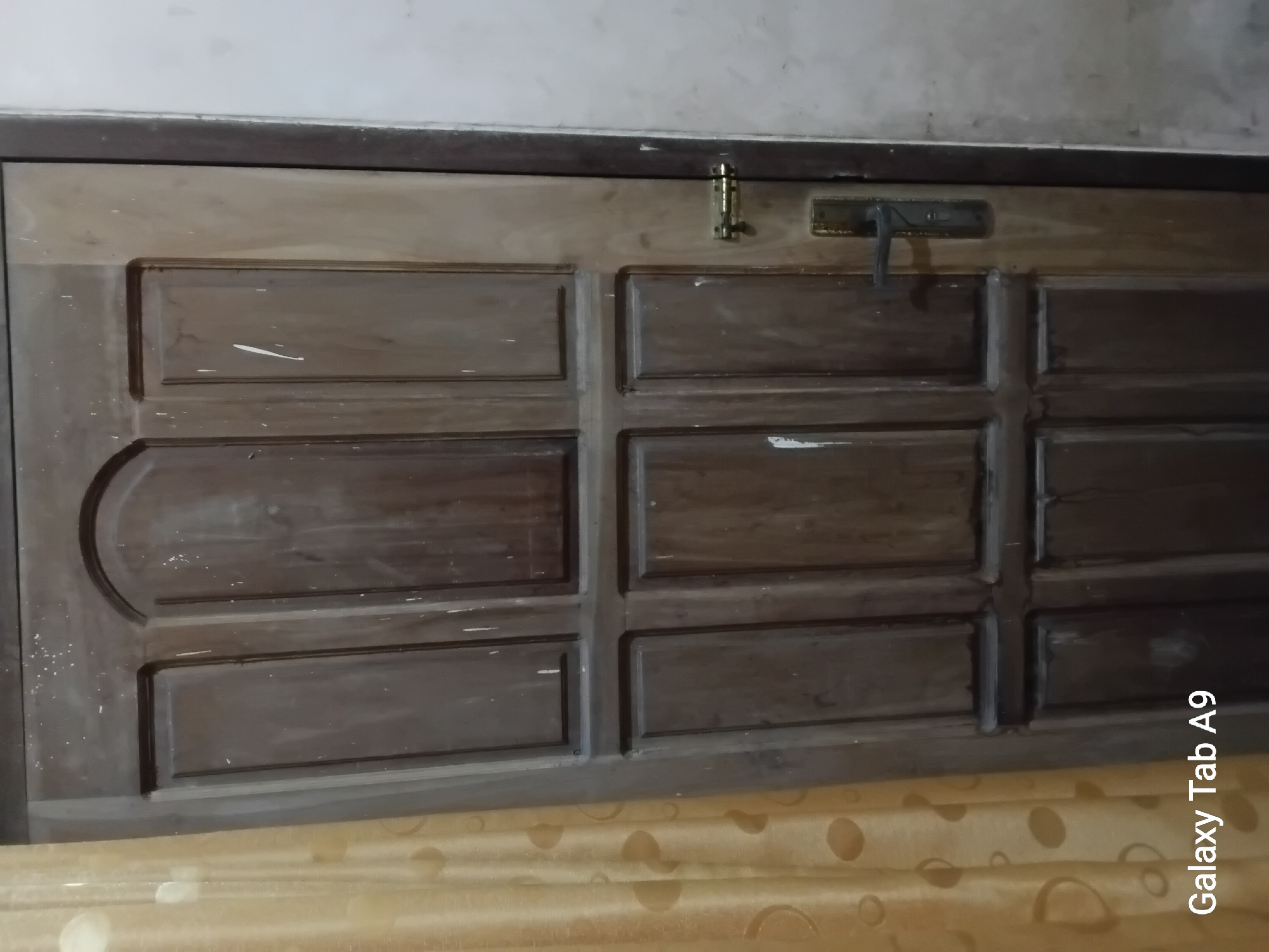 An exterior view of a dark brown wooden door with several panels is captured in the image. The door has a brass doorknob and lock mechanism visible on the right side. The door appears to be in a residential setting, with a yellow curtain visible to the left and a light-colored wall to the right. There are white smudges or paint marks on the surface of the door, suggesting it may be old or in need of maintenance. The image is stamped with "Galaxy Tab A9" at the bottom left corner. The location is the city of Trenggalek, Indonesia.