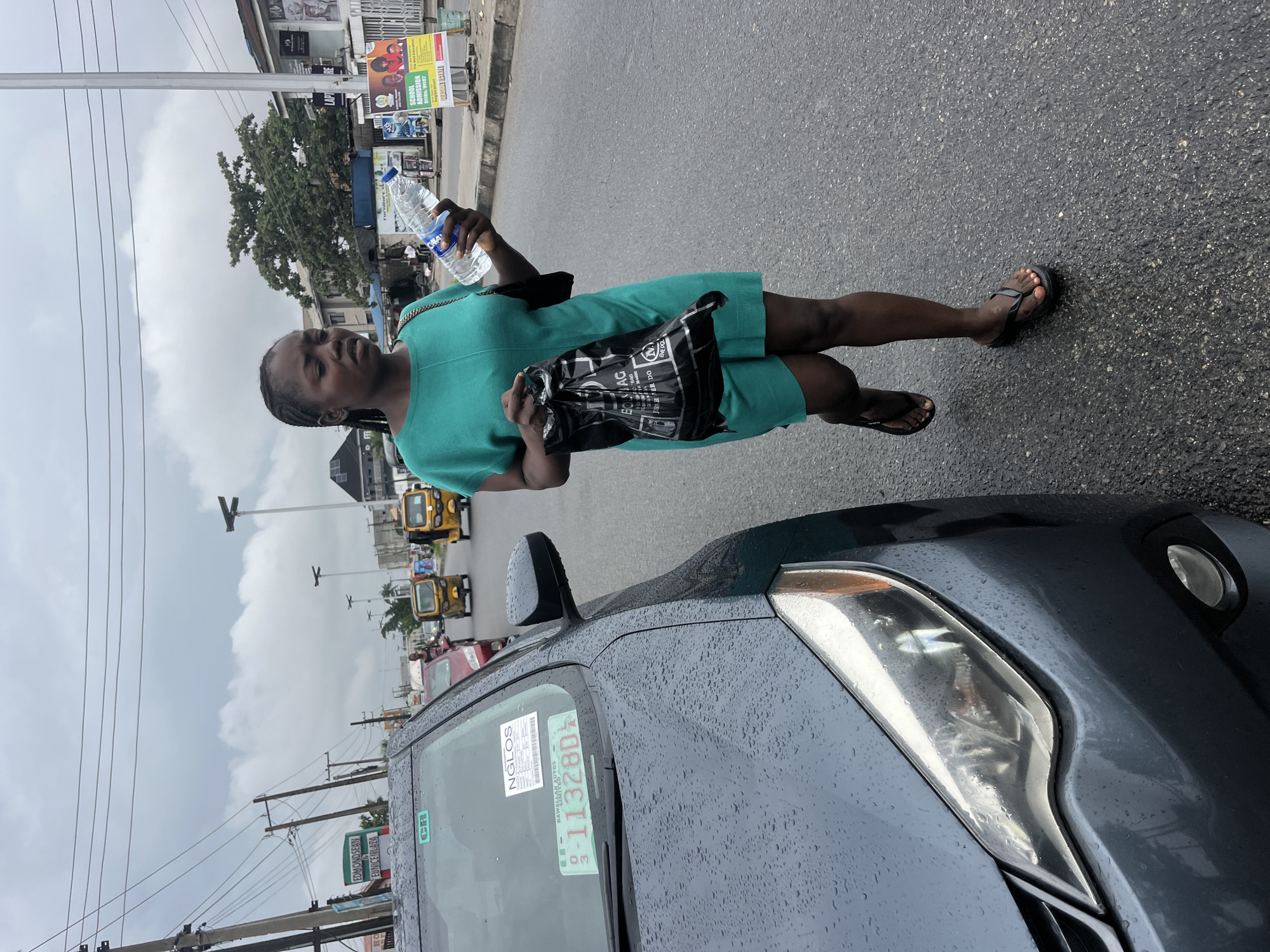 In Ado, Nigeria, a young woman in a teal dress walks on a wet, asphalt road, holding a plastic water bottle in her right hand and a black plastic bag in her left. A dark gray car, speckled with water droplets, is parked to her left. In the background, yellow auto-rickshaws are visible, along with utility poles and a cloudy sky. The scene suggests a bustling urban environment, possibly after rainfall.