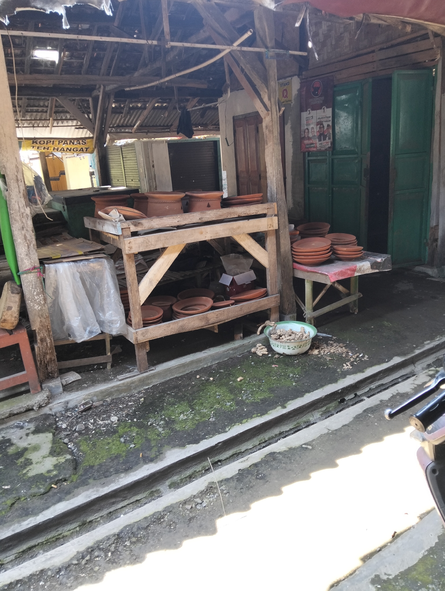 The image depicts an outdoor marketplace stall in Muncar, Indonesia. The stall, constructed from weathered wood and corrugated metal roofing, displays an array of terracotta pottery, including plates and large vessels. A sign advertising "KOPI PANAS TEH HANGAT" (Hot Coffee, Warm Tea) hangs within the stall. Adjacent to the stall, a green door leads into a building. In the foreground, a shallow concrete drainage channel runs alongside the pathway, which is partially covered in moss and debris. A metal bowl filled with what appears to be ginger is placed on the ground near the stall. The overall scene suggests a humble, functional marketplace environment.