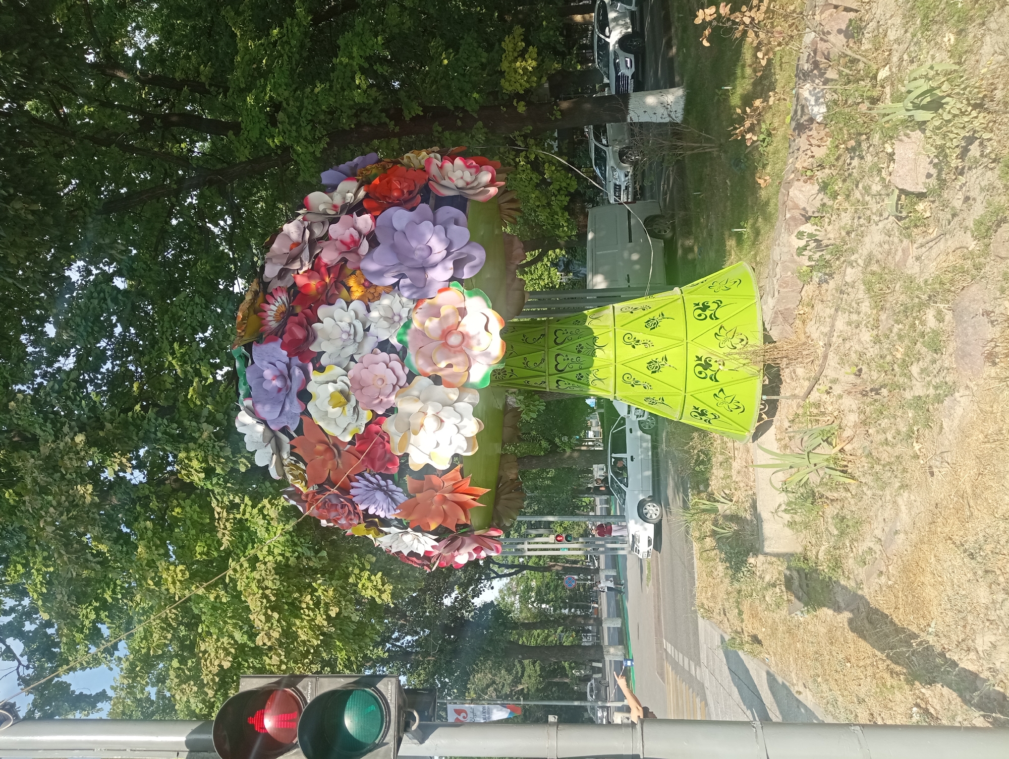 A vibrant, oversized floral sculpture, resembling a bouquet of paper flowers atop a green conical base with decorative cutouts, stands on a patch of dry grass and sparse vegetation near a city street. To the left, a traffic light shows a red pedestrian signal and a green vehicle signal. Several cars are visible on the street, including white sedans and SUVs. In the background, mature trees with dense green foliage line the street, and buildings are partially visible. The scene appears to be an urban environment, possibly a park or public square, located in Shymkent, Kazakhstan. The overall lighting suggests a sunny day.