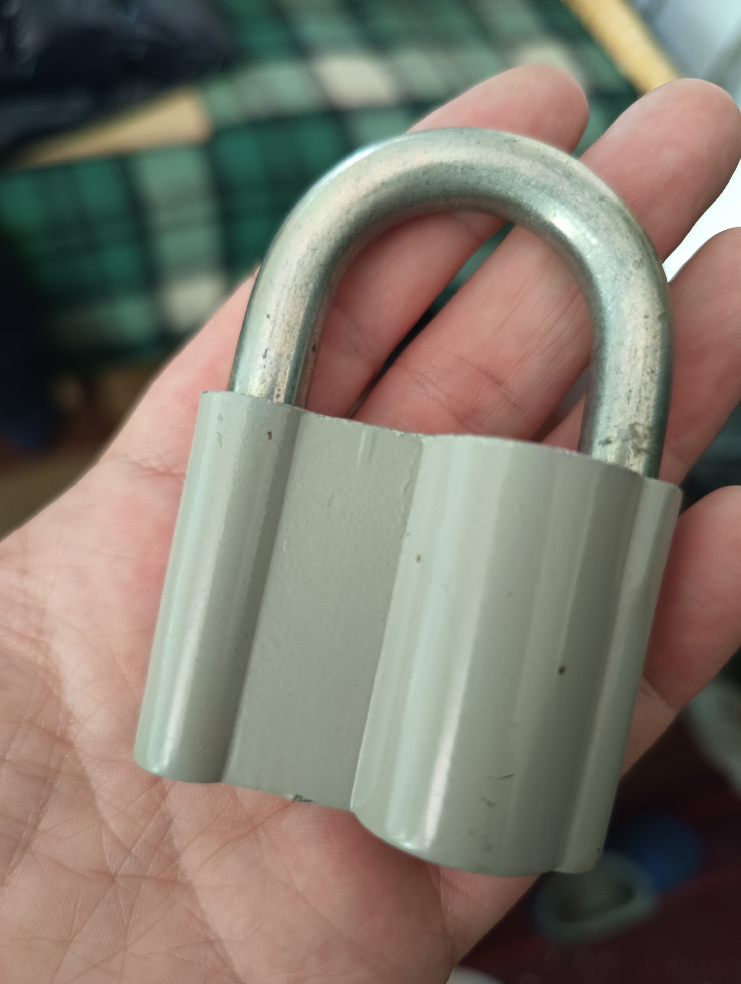 A close-up image features a human hand holding a grey padlock against a blurred background. The hand, with visible skin texture and creases, supports the base of the padlock. The padlock itself has a sturdy, light grey body with a slightly ribbed or fluted design running vertically along its sides. The metallic shackle, the U-shaped component at the top, is silver-colored and shows minor signs of wear, including scuffs and small dark specks. Similar small imperfections are also present on the grey body of the lock.
The background is softly out of focus, displaying indistinct patterns in shades of green, with hints of white and yellow, suggesting a textile like a blanket or upholstery in an indoor setting. The lighting is soft and diffused, making the specific time of day indeterminate. No particular activity beyond holding the padlock is depicted, and no text is visible on the padlock or in the background. The image is associated with the location context of Biliaivka, Ukraine, although this specific geographical location is not visually evident within the frame.