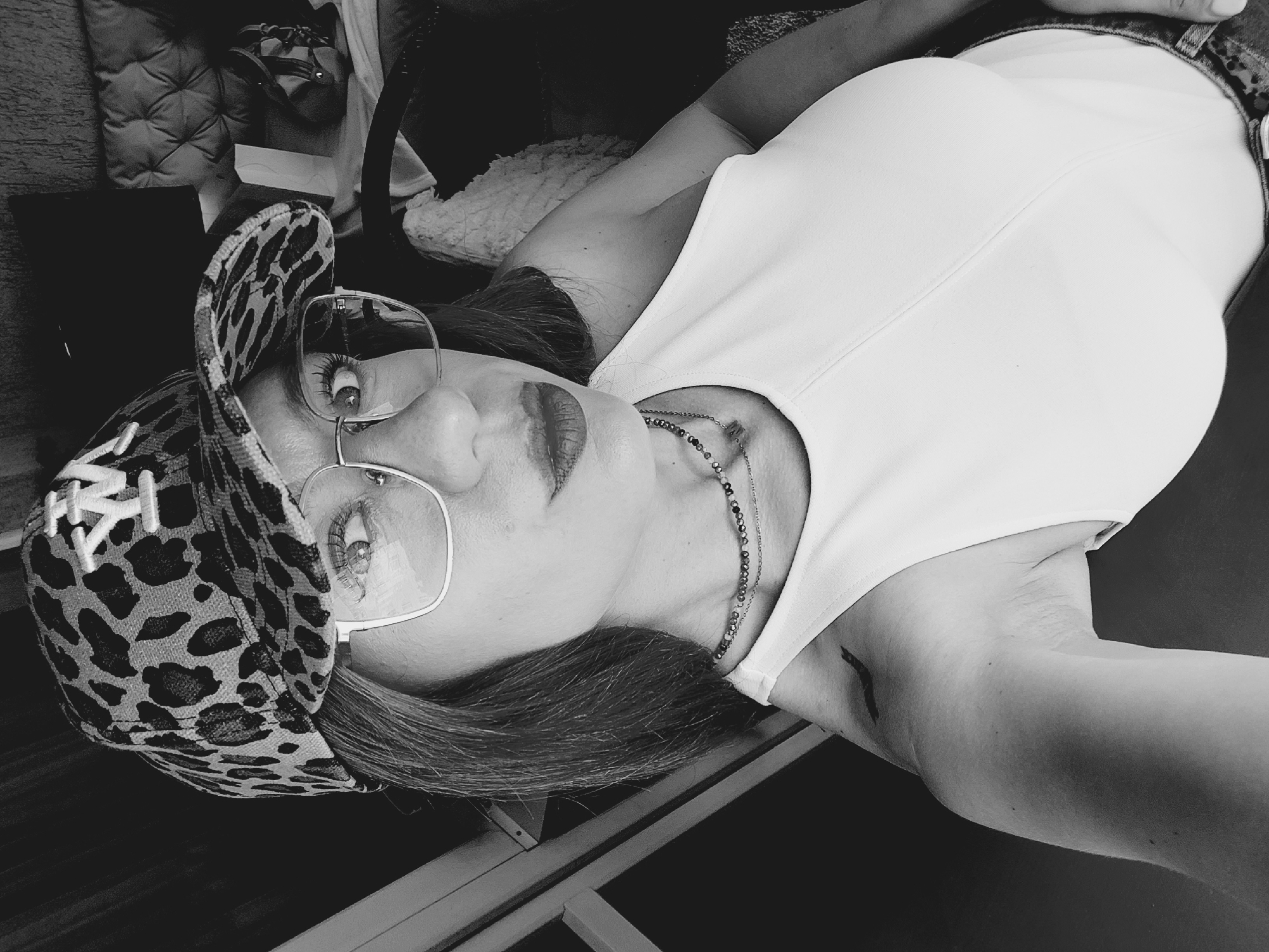 A black and white, high-angle selfie shows a woman wearing a leopard-print baseball cap with a white 'NY' logo, and large, square-rimmed glasses. She has dark lipstick and is wearing a white tank top and a beaded necklace. A faint tattoo is visible on her upper left arm. She is looking directly at the camera with a slight pout. The background is blurry but suggests an indoor setting with furniture and possibly a window. The image does not provide any information about the location in Sofia, Bulgaria.