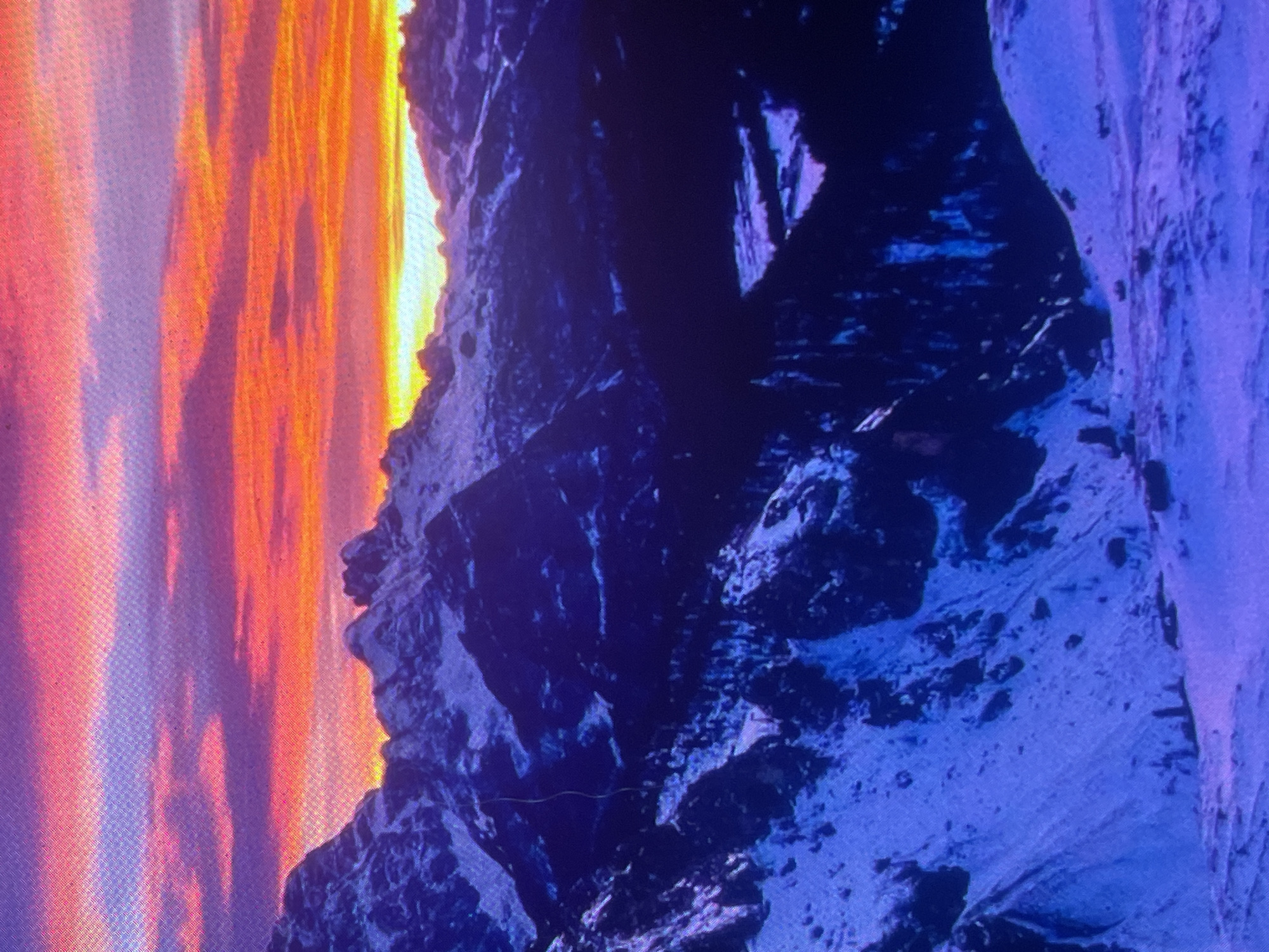 The image depicts a high-altitude, snow-covered mountain range under a dramatic sunrise or sunset, exhibiting a visible pixel grid consistent with a photograph of a digital screen. The sky transitions from soft pink and purple clouds at the top to intense horizontal bands of orange, red, and yellow nearer the horizon, where a bright yellow-white glow indicates the sun's position. Jagged, snow-capped mountain peaks, rendered in deep blues and purples by the ambient light, dominate the midground and background, casting deep shadows into vast, dark valleys dense with what appear to be coniferous trees. A more open, possibly cleared valley floor is faintly visible on the midground right. The foreground is a wide expanse of undulating snow, also depicted in shades of blue and violet. No discernible people, specific artificial structures, or wildlife are present. This high-alpine, snowy mountain landscape is geographically inconsistent with the typical characteristics of settlements named Zelenyi Hai in Ukraine, which are generally situated in low-lying agricultural or forested plains; therefore, the scene depicted cannot be located within Zelenyi Hai, Ukraine.