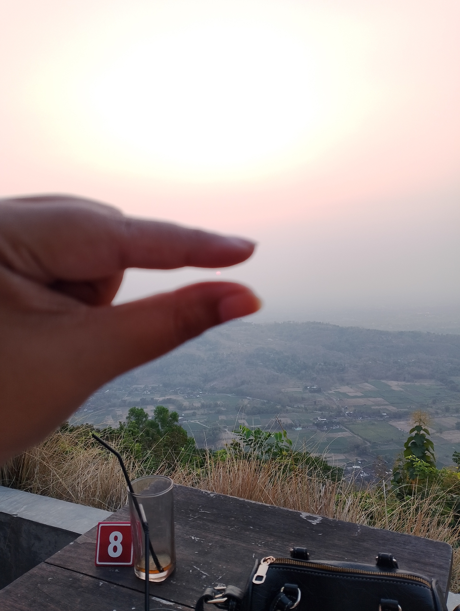 An outdoor shot captures a person's hand in the foreground, fingers pinched to create a small gap through which a distant, hazy sunset or sunrise is visible. The hand is blurred, suggesting it's close to the camera. Below the hand, a wooden table is set with a glass containing two black straws and some liquid, and a red table number '8'. Behind the table, dry, tall grass grows on a slope, and beyond that, a vast valley unfolds. The valley is dotted with fields, some appearing cultivated, and scattered buildings, all softened by a hazy atmosphere. Trees and hills are visible in the midground and background, merging into a pale, bright sky. The location is identified as Srimulyo, Indonesia, implying this is a scenic viewpoint overlooking the landscape.