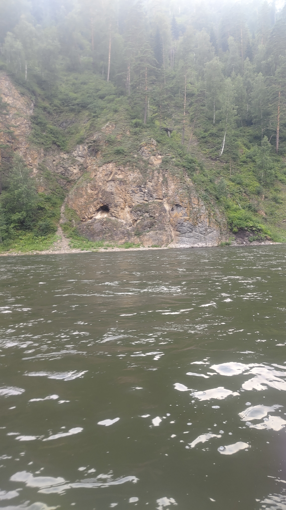 The image shows a wide river flowing past a steep, rocky cliff face covered in dense green forest. The cliff has several caves or openings visible. The water is choppy with white foam and reflections from the sky. The sky is overcast and hazy, obscuring the tops of the trees and the upper reaches of the cliff. The overall scene suggests a natural, remote environment, possibly a natural park or wilderness area. The location is the Mana River in the vicinity of Ust'-Mana, Krasnoyarsk Krai, Russia.
