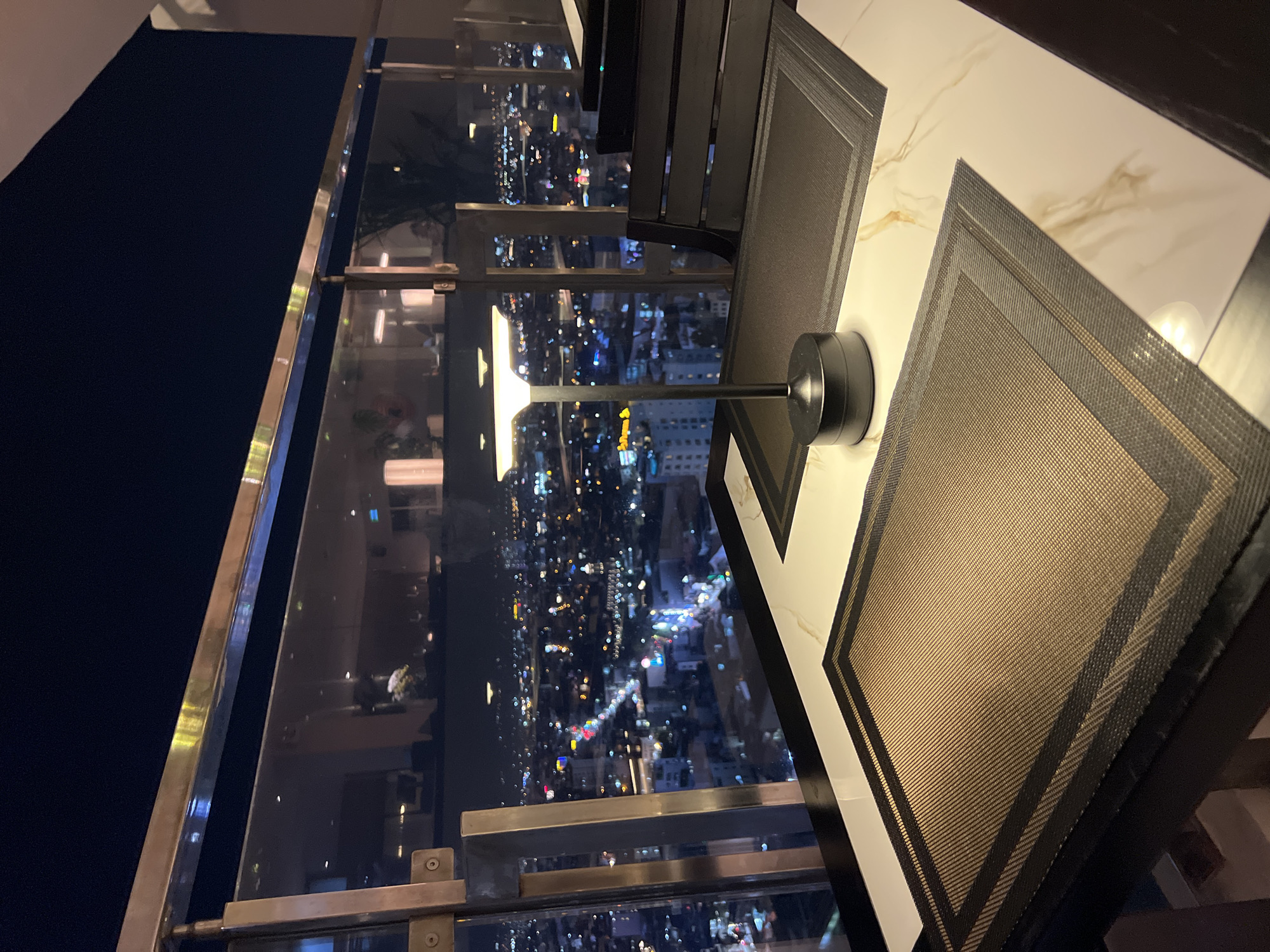 Here is a forensic description of the image:
The scene is indoors, seemingly in a high-rise building, overlooking a densely populated cityscape at night. The location is indicated as Nha Trang Ward, Vietnam, based on the provided information.
Visible through a large glass window with a metallic frame is a sprawling urban area illuminated with numerous lights, likely from buildings and streets. The city extends into the distance. The perspective suggests a high vantage point, likely from an upper floor.
Inside, on a marble or similar countertop, are two placemats with a dark border. A desk lamp with a small, modern design is on the countertop, close to the window. There's a chair partially visible in the background.
The focus is on the contrast between the brightly lit cityscape outside the window and the interior setting with placemats and a lamp. There are no people visible in the image.