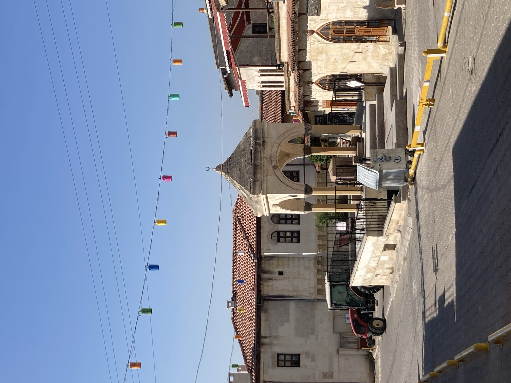 The image depicts a street scene in Avanos İlçe Merkezi, Turkey. In the foreground, a cobblestone street is lined with yellow and white traffic barriers. A red tractor is parked on the side of the street. In the center of the image, there is a small, ornate stone structure with arched openings. Beyond this structure, buildings with red-tiled roofs are visible. Colorful lanterns hang from wires stretched across the sky. The sky is clear and blue. The overall impression is one of a traditional Turkish town on a bright, sunny day.