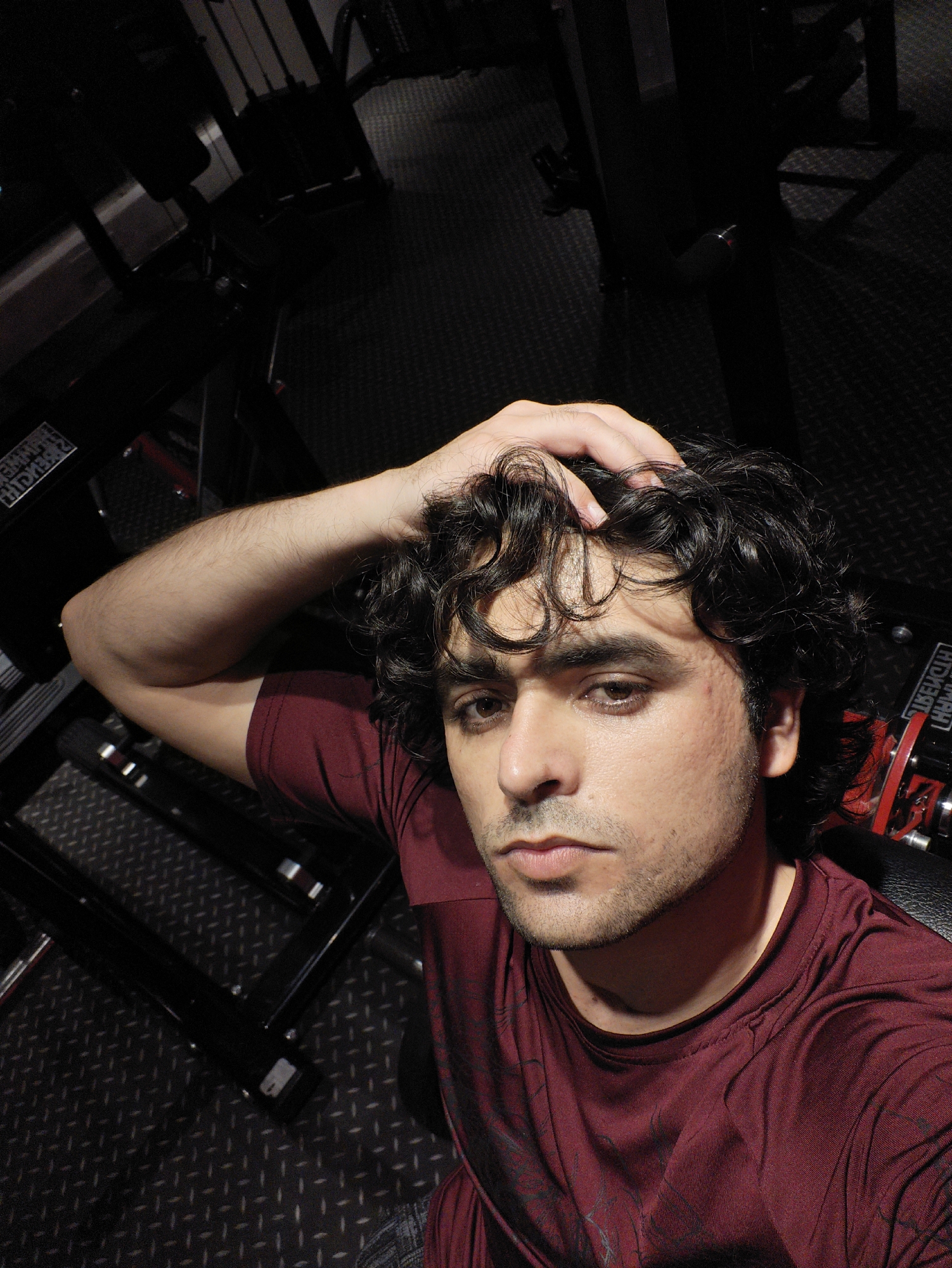 A man is pictured in what appears to be a gym. He has dark, curly hair and is wearing a maroon t-shirt. His right arm is raised, with his hand touching his head. The background features exercise equipment and a textured floor, typical of a gym setting. No features indicate the location in Chongqing High-tech Zone, China.