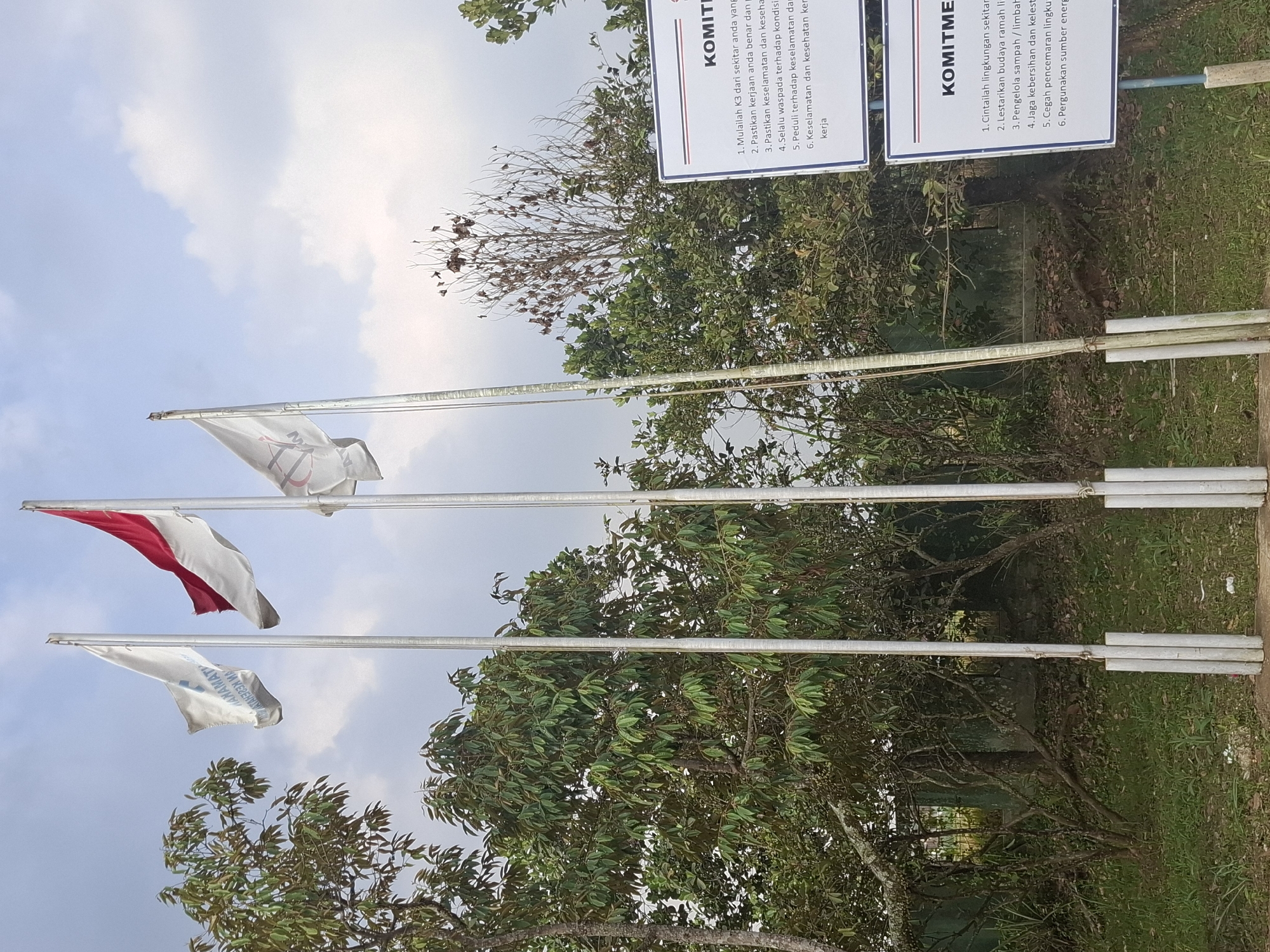 The image shows three flagpoles with flags in front of a background of trees and sky. One flag is red and white, the national flag of Indonesia. Another flag is white with blue text, and the third flag is white with a red and black logo. Two signs with text in Indonesian are visible in the foreground. The sign on the right has the title "KOMITME" followed by six numbered points. The sign on the left also has the title "KOMITME" followed by six numbered points. The scene appears to be outdoors, possibly in a park or public area. The sky is partly cloudy. The location is identified as Ciheulang Tonggoh, Indonesia.