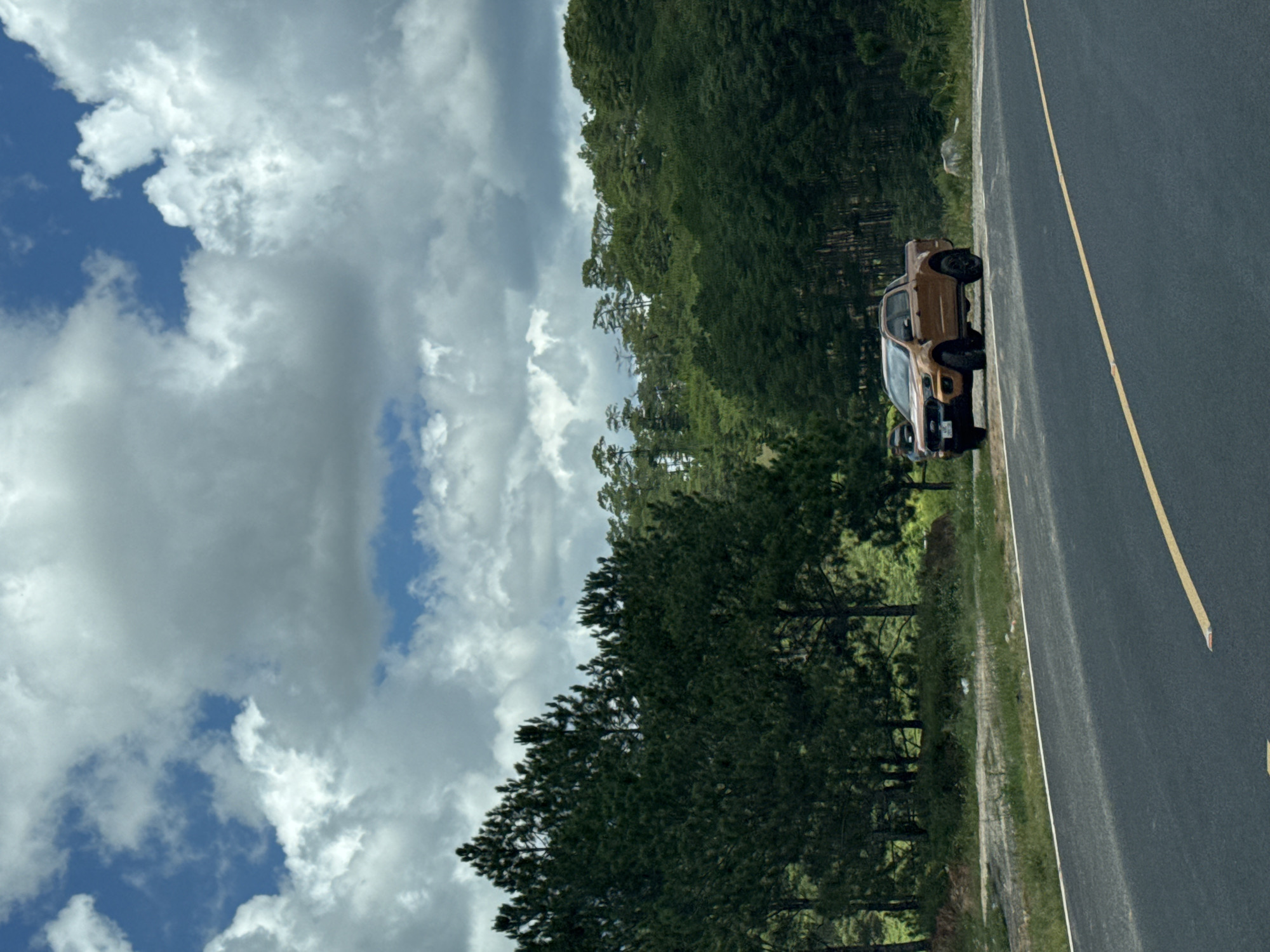 The image depicts a brown Ford Ranger pickup truck parked on the shoulder of a paved road. The truck is positioned on the right side of the frame, facing left. The road has a yellow lane divider. In the background, a dense forest with tall pine trees extends up a hillside. The sky is partly cloudy with large, white cumulus clouds against a blue backdrop. The location appears to be a rural or mountainous area, possibly on the outskirts of Đa Bla, Vietnam, given the presence of pine trees common in the Central Highlands region. There are no people clearly visible in the immediate vicinity of the truck. The overall scene suggests a stop along a scenic route.