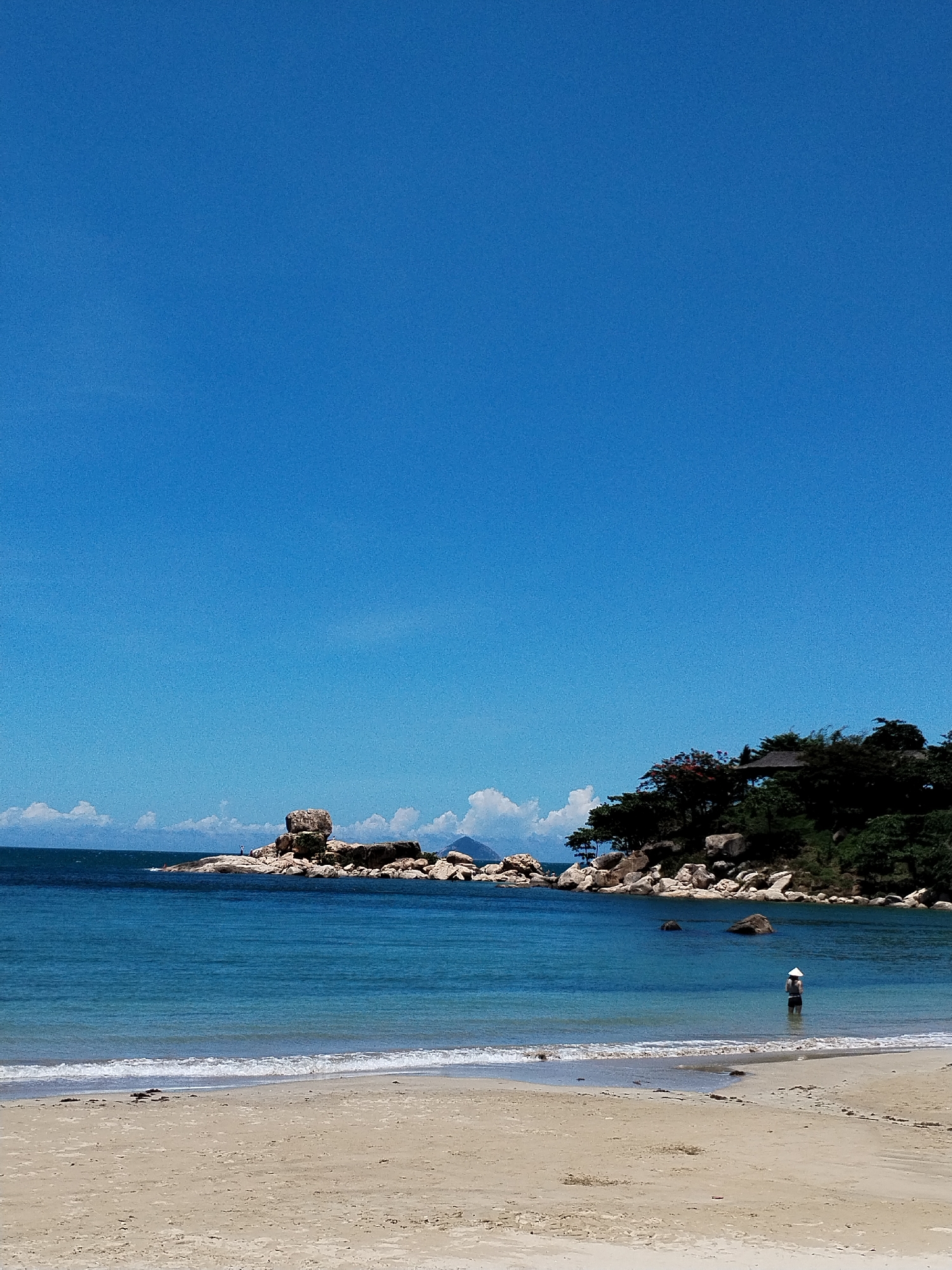 The image shows a beach scene in Bac Nha Trang Ward, Vietnam. The sky is clear and blue. A body of water extends towards a rocky outcrop with trees on the right. A person wearing a conical hat stands in the shallow water near the shoreline. The beach has light tan sand.