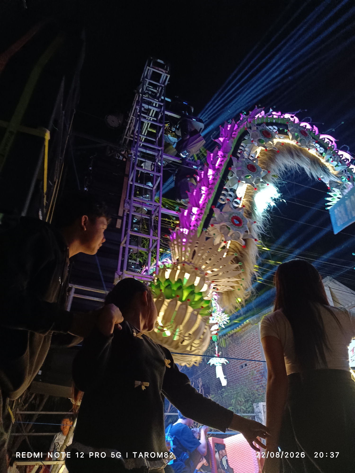 Three individuals are observing a colorful dragon float, illuminated by purple and white lights, at night. The float is elaborate, featuring textured elements and appears to be suspended or mounted on a structure. Behind the float, stage lighting equipment is visible, with blue light beams projecting upwards. The scene is set outdoors, with brick structures and foliage in the background. The timestamp "22/08/2026 20:37" and the text "REDMI NOTE 12 PRO 5G | TAROM82" are overlaid on the image. The location is specified as Mundusewu, Indonesia.