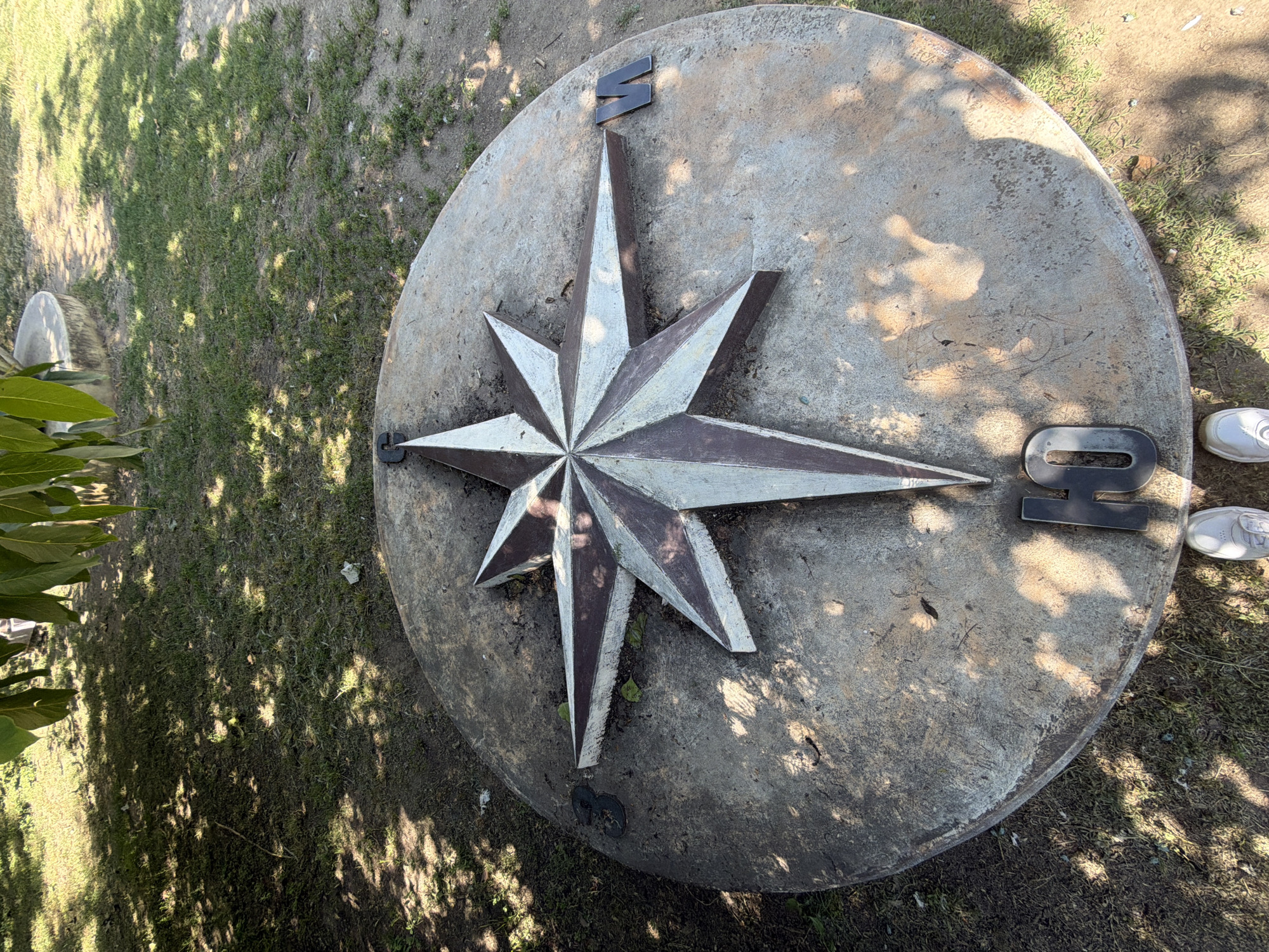 The image shows a large, circular concrete compass rose embedded in the ground in an outdoor setting. The compass rose has a prominent eight-pointed star in its center with alternating dark brown and light grey/white segments. The cardinal directions are marked with letters: "W" for West on the right side of the compass, and "S" (South) represented by the Cyrillic letter "Ю". The compass is weathered and shows signs of age and dirt.
To the lower center of the image, the feet of a person wearing white sneakers are visible, suggesting the photo was taken from a standing or slightly elevated perspective. The ground surrounding the compass is covered with grass and dirt, with fallen leaves and shadows from overhead trees creating a dappled pattern. In the background, out of focus, are more trees and possibly some concrete structures.
The specific location of this compass rose is in the **Central City Park of Pazardzhik, Bulgaria**. This compass rose is a well-known landmark situated near the main entrance of the park, close to the children's playground and the outdoor stage.