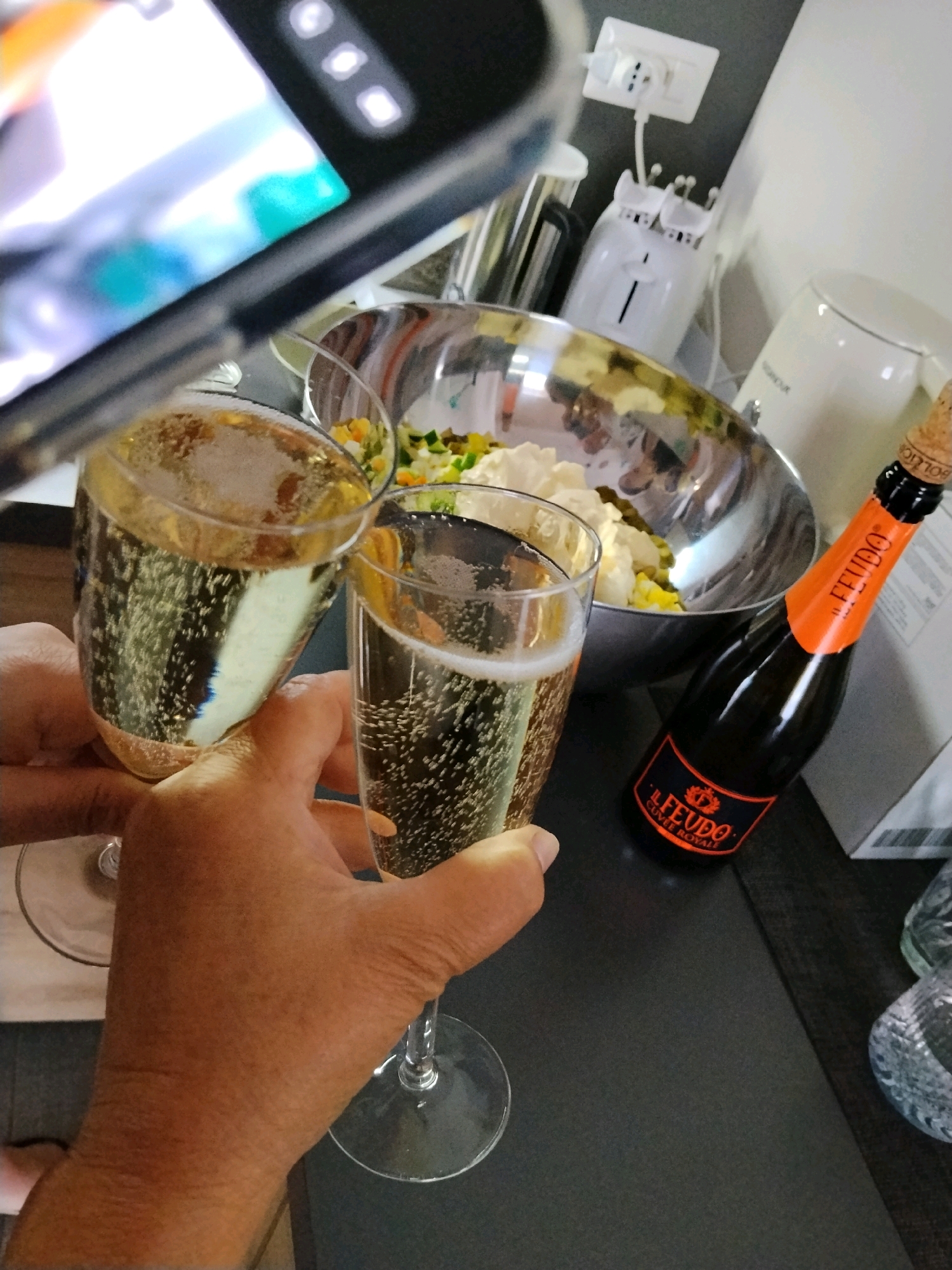The image depicts a close-up, slightly angled view of two glasses filled with a sparkling beverage, likely champagne or prosecco, being raised in a toast. Bubbles are visible within the liquid. In the background, a silver mixing bowl contains what appears to be a chopped salad or vegetable mix. To the right of the bowl, a small, dark bottle with an orange label, identified as "IL FEUDO Cuvée Royale," is lying on its side. The bottle has a cork and wire cage. A white electrical outlet with a plug connected to an appliance is visible on the wall behind the bowl. The overall setting appears to be an indoor kitchen or dining area, with a dark countertop surface in the foreground. The image is taken from the perspective of someone holding a smartphone, with the top portion of the phone and its screen visible in the upper left corner, suggesting the photo is being taken with the phone. No specific location in Bitonto, Italy, can be identified from the visual information.