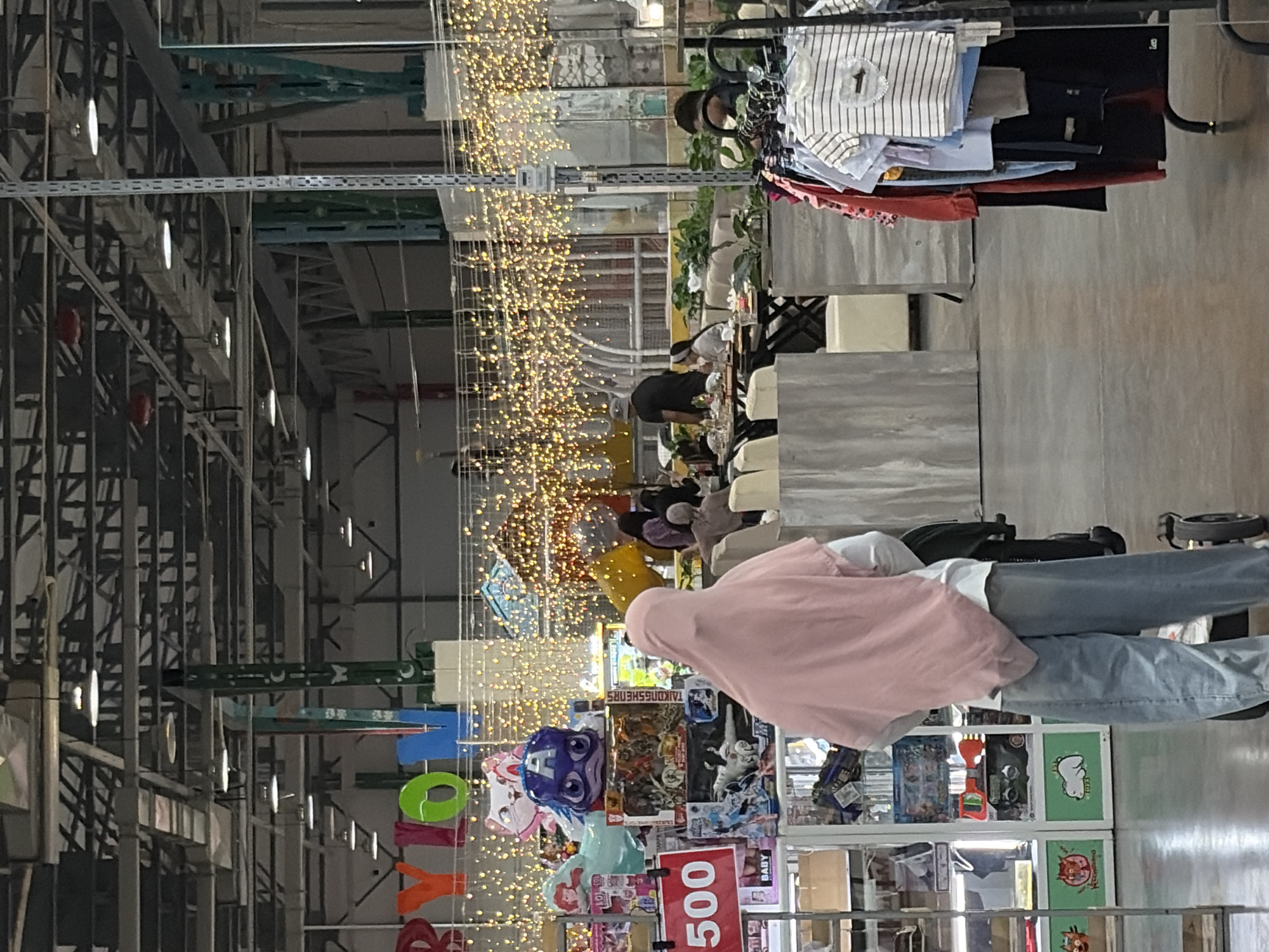 The image depicts an indoor scene, likely a market or store, with numerous people present. The setting appears to be a large, open space with a high ceiling, characterized by industrial-style metal shelving and overhead lighting. Strands of small, yellow lights are strung across the space, creating a festive or decorative atmosphere.
Several individuals are visible, some standing, others seated. One person in the foreground is wearing a pink hooded garment and blue jeans, pushing a trolley. Other people are browsing or interacting in the background, where tables and chairs suggest a seating area. The overall impression is one of a busy commercial environment.
Objects in the scene include clothing displayed on racks, various products on shelves, and decorative items like balloons and the string lights. Some of the products appear to be toys or children's items, judging by the colorful packaging and characters visible. There is also a red sign with white text that reads "500". The time of day is not explicitly indicated, but the indoor lighting suggests it could be day or evening. No specific weather conditions can be inferred from the image. The provided location context is Kant, Kyrgyzstan.