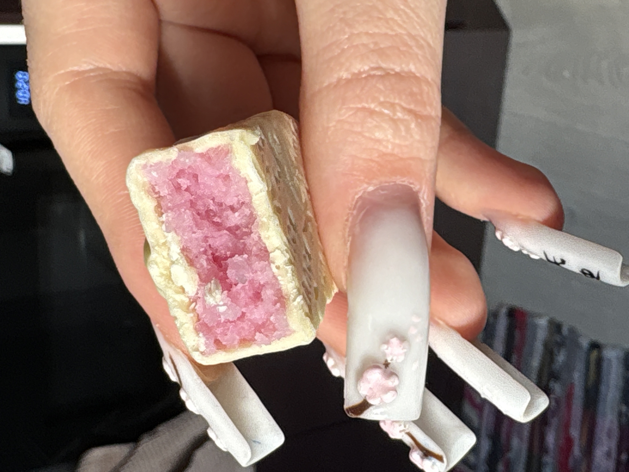 In the city of Chaikovsky, Russia, a close-up image displays a fair-skinned human hand holding a small, rectangular confectionery item. The confectionery is approximately 2-3 cm in length and has been broken, exposing a vibrant pink, crystalline or crumbly interior filling. This filling is contained within a pale yellow or cream-colored outer layer. The hand features long, white, squared artificial nails; two nails exhibit three-dimensional pink floral designs resembling cherry blossoms, and one nail has faint black numerical or symbolic markings. The background is dark and out of focus, with a digital display showing "08:30" discernible in the upper right portion of the frame, indicating an indoor environment. No other individuals or specific environmental features are identifiable.