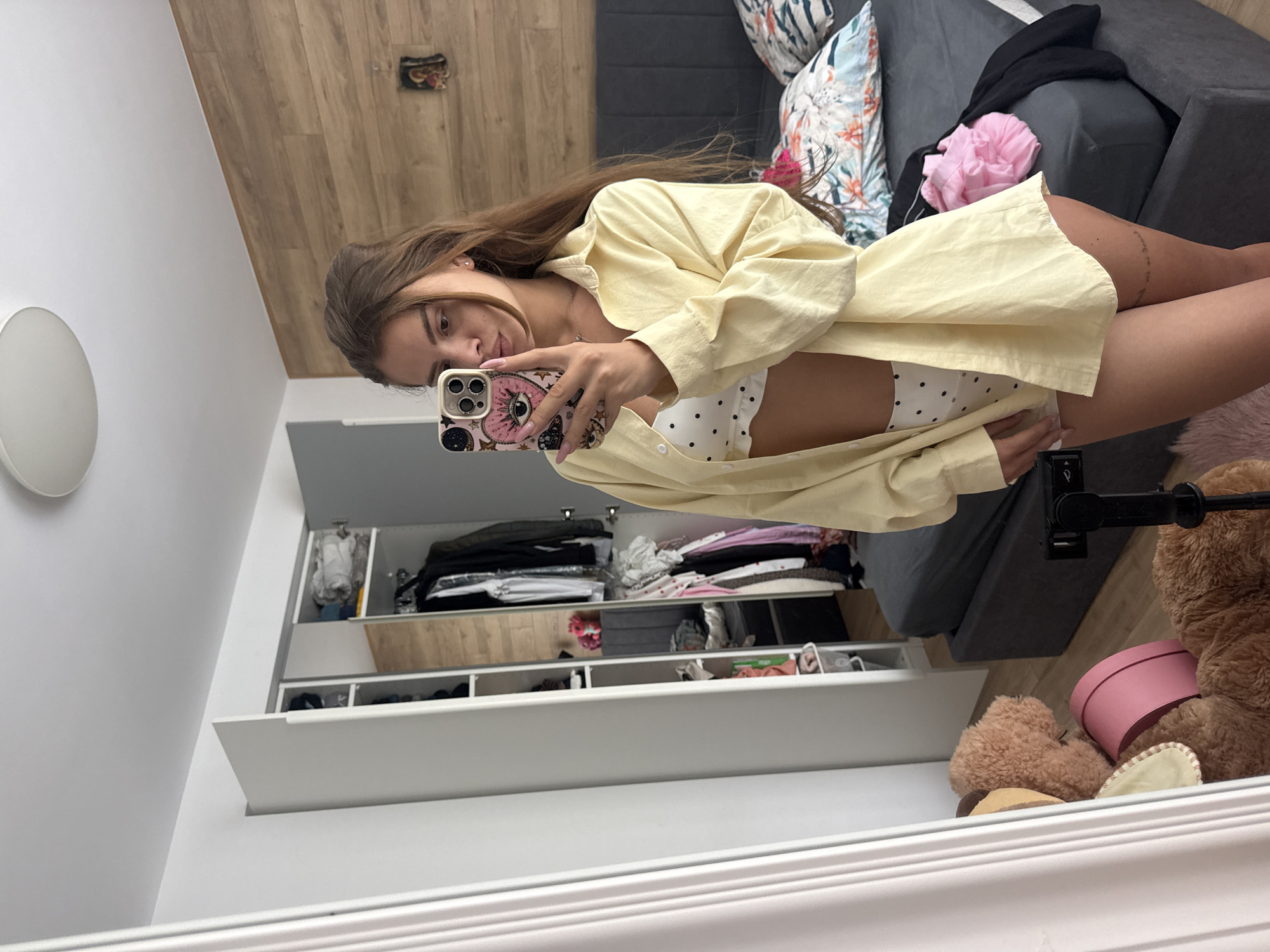 A young woman with long brown hair takes a selfie in a bedroom. She is wearing a yellow unbuttoned shirt over a white polka dot bralette and matching underwear. The room features a large mirror on the left, a wardrobe with clothes inside on the left, a bed with a grey headboard and floral pillows on the right, and a wooden-paneled wall behind the bed. A teddy bear and a pink hatbox are visible on the floor in front of the mirror. A tripod with a camera is positioned in the foreground. The location is not specified, and no information regarding Plovdiv, Bulgaria is present in the image.