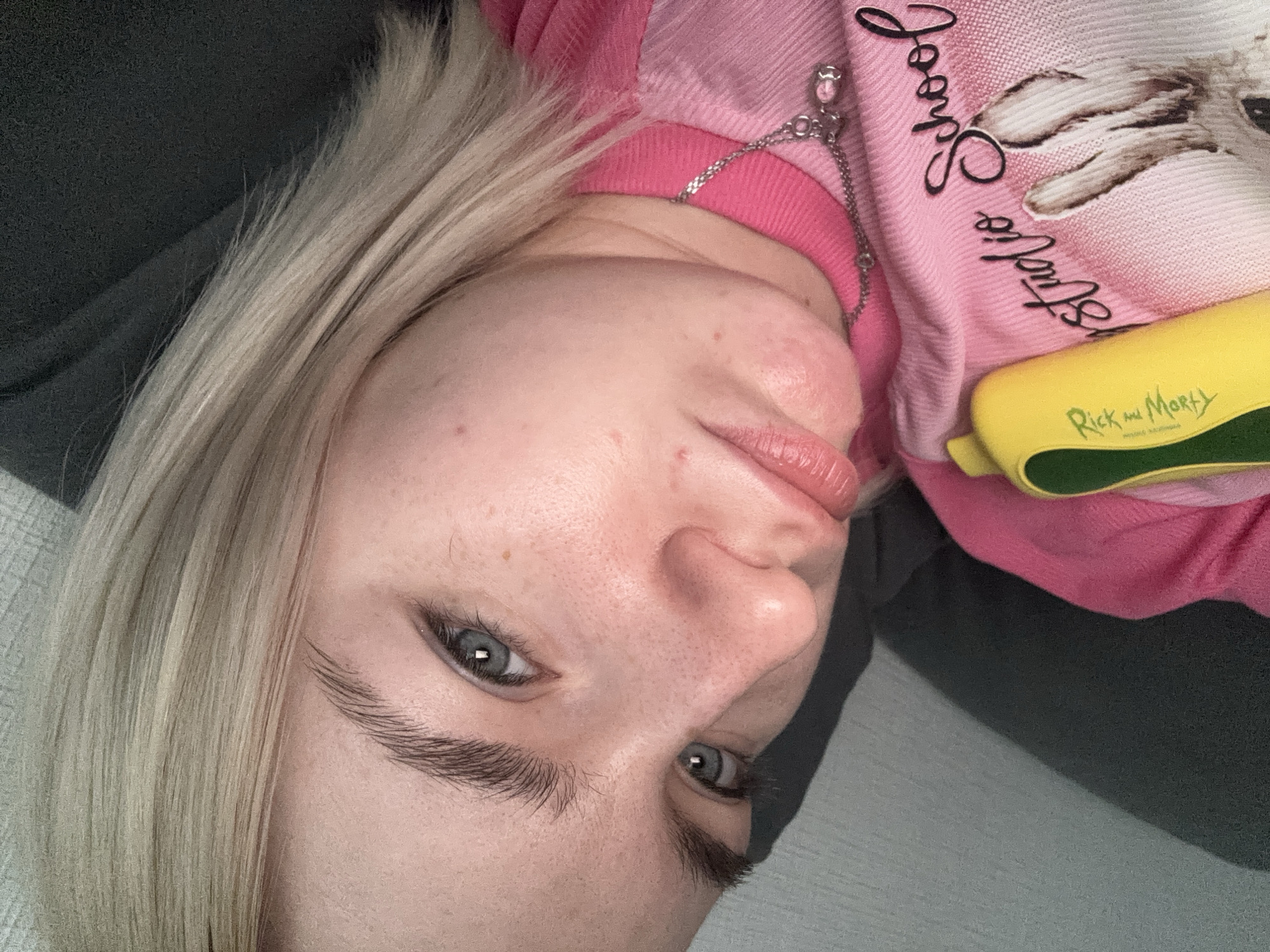 A close-up, rotated view shows a fair-skinned person with light blonde hair. The person is wearing a pink sweatshirt featuring an image of a white animal paw and the words "Looney Studios". A silver chain necklace is visible around their neck. A yellow and green object with "Rick and Morty" printed on it is also in the shot. The background includes a dark gray surface and a light gray surface. The location and city in Russia are unknown.