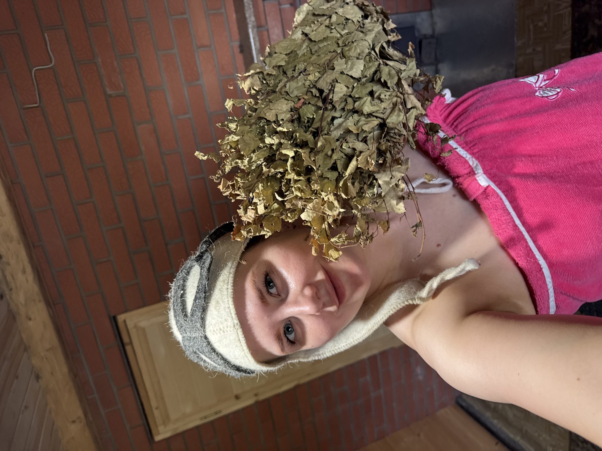 A rotated, close-up shot shows a woman in what appears to be a sauna. She is wearing a grey and white striped sauna hat and a pink towel dress with white trim. A dried birch broom sits atop her head. The sauna walls are brick, and a wooden door is visible on the left. The city and specific location are not discernible from the image.