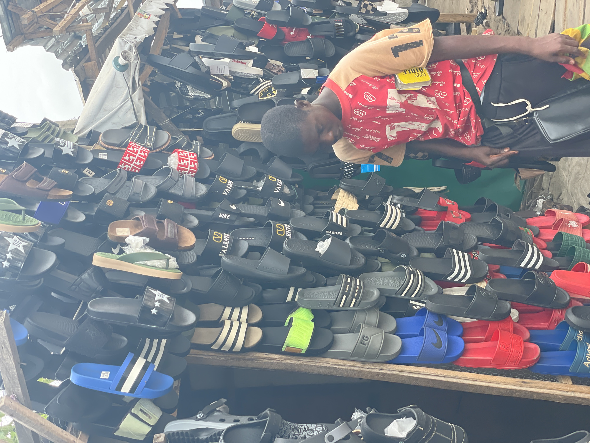 This image captures an outdoor market stall in Badagry, Nigeria, bustling with a vast display of footwear. The central subject is a young male vendor, leaning over the merchandise, appearing to organize or tend to the shoes. He wears a red and beige t-shirt with stylized "Balenciaga" text, carries a black shoulder bag, and has a yellow "Good News Bible" tucked into his side. He holds a colorful red, yellow, and green rag in his right hand.
The setting is a makeshift market stall, constructed from wooden planks and a corrugated metal sheet or tarp, with a light fixture visible overhead. The stall is overflowing with numerous pairs of slide sandals and flip-flops, stacked high on shelves. The footwear exhibits a wide range of designs and colors, many of which mimic popular brands. Visible brand names and text on the shoes include "Adidas," "Nike," "LOUIS VUITTON," "BALENCIAGA," "VALENTI," "VALD," "Timberland," "FG," "LUORU," "WASONS," "SAOMA," and "DG." The text "GOOD NEWS BIBLE" is clearly visible on the vendor's Bible. The scene takes place during the daytime, under what appears to be an overcast sky or within a shaded area of the market, highlighting a typical informal trading environment.