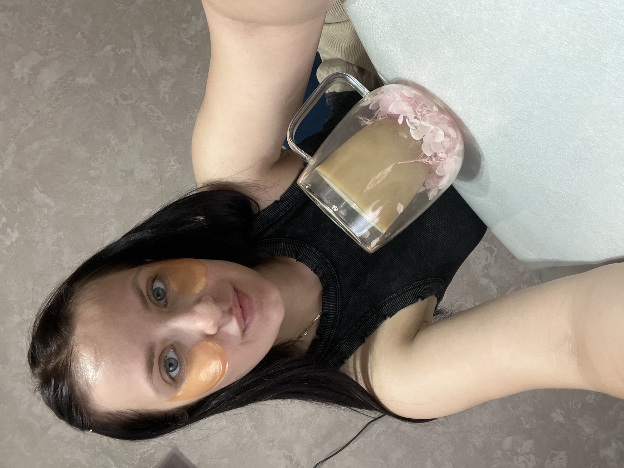 A person lies on a gray floor and white furniture, holding a glass mug with a brown liquid and pink flowers. The person has black hair and is wearing orange eye patches. They have fair skin and blue eyes. They are wearing a black top and a gold necklace. A wire is visible next to the person. The location may be in the city of Марковский, Russia, but this is unconfirmed and only based on the provided information.