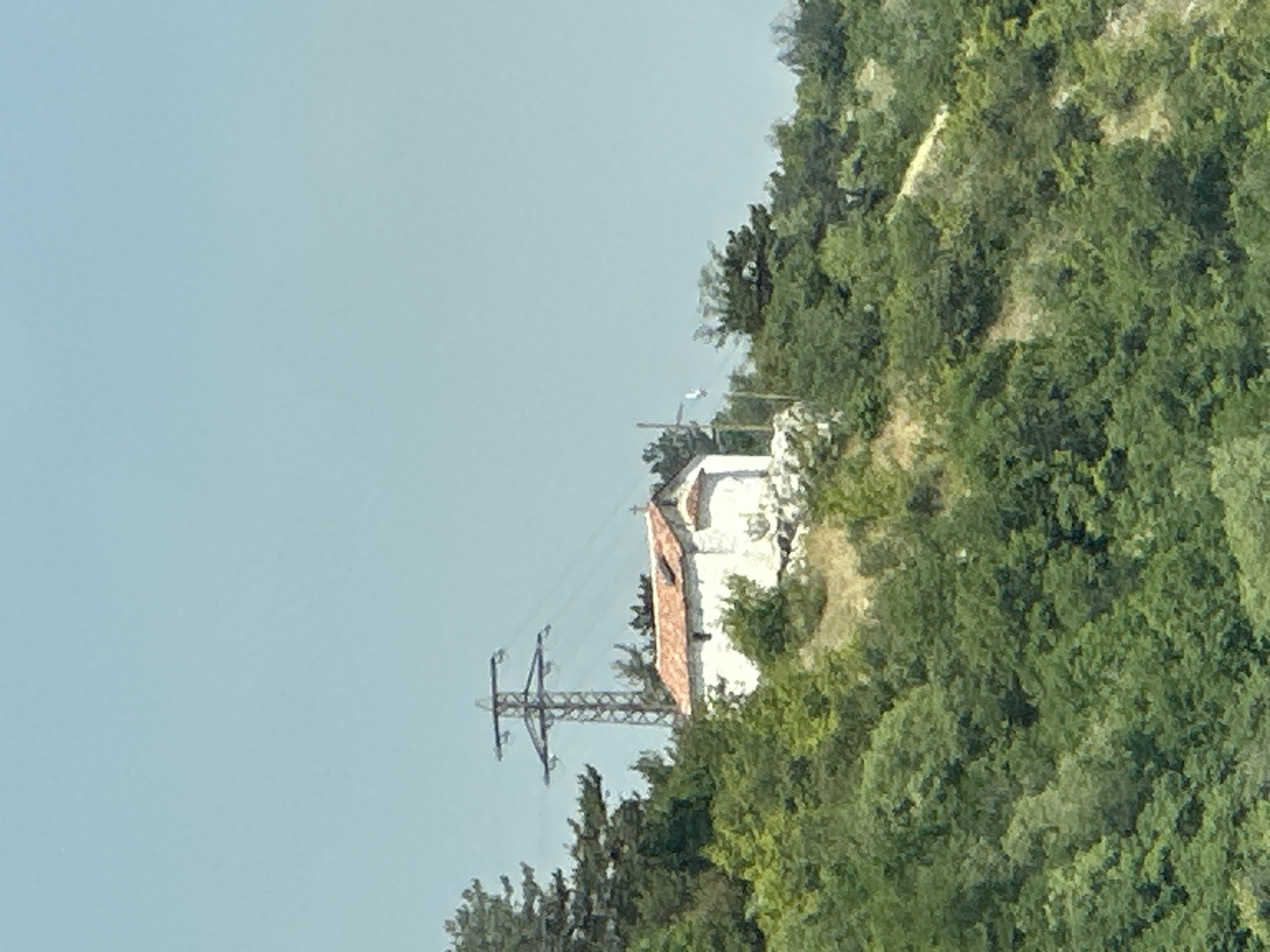 This image depicts a hilly, forested landscape under a clear, pale blue sky, located in Asenovgrad, Bulgaria. A small, single-story, white stucco building with a gabled roof covered in reddish-orange tiles is centrally positioned on a rocky, elevated outcrop. To the immediate right of the building, a thin white pole with an indistinct top element is visible. Behind and slightly to the left of the building, a tall metal lattice power transmission tower, equipped with multiple horizontal cross arms and power lines, stands prominently. The slopes are densely covered with various green trees and shrubs, interspersed with patches of lighter-colored, dry earth. No human figures or animals are present in the scene.