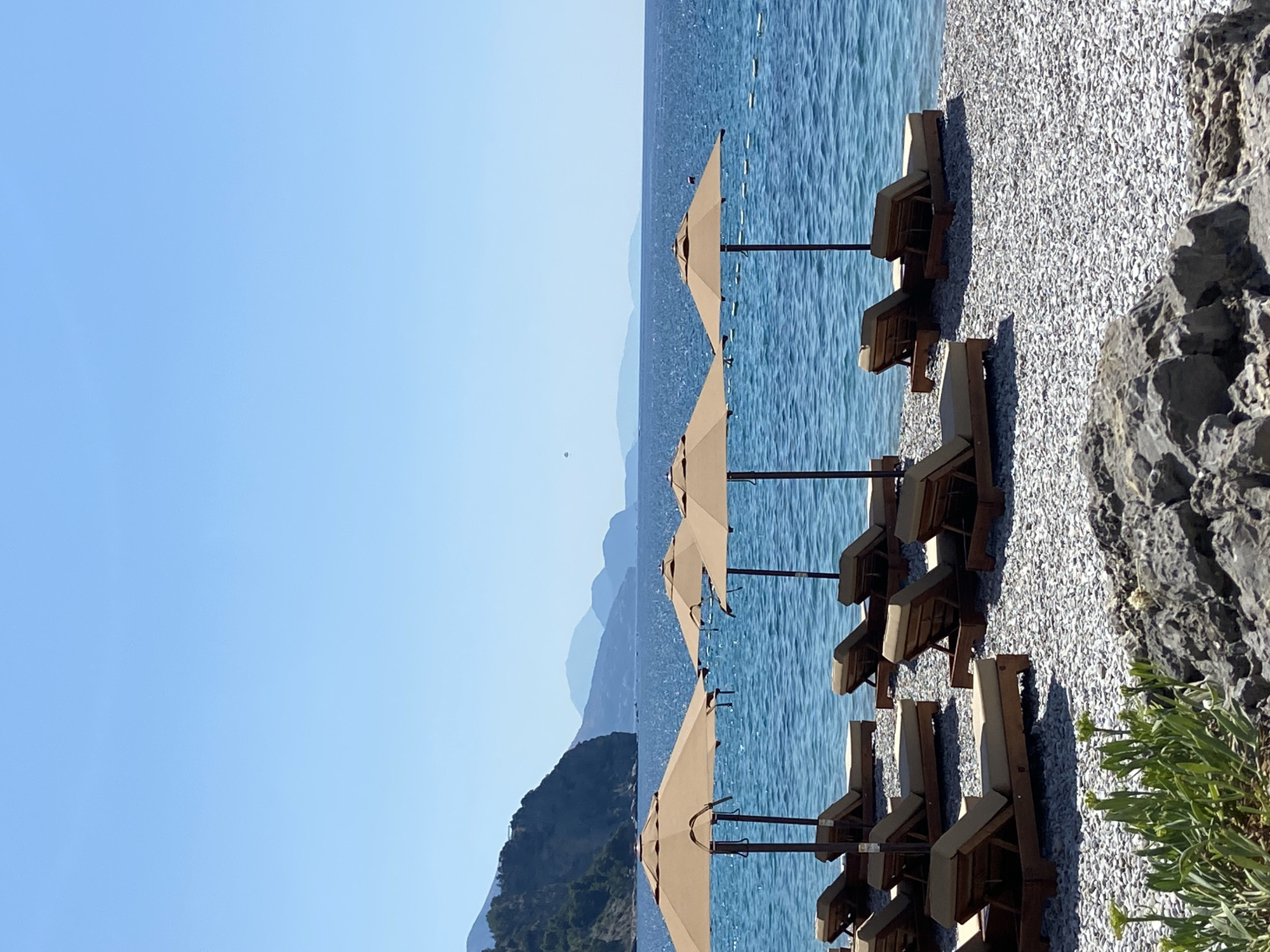 This is a sunny, eye-level shot of a beach in Prijevor, Montenegro. The foreground features a rocky shoreline with green plants. In the middle ground, rows of empty, tan-colored lounge chairs and umbrellas are set up on a pebble beach. The umbrellas are spaced evenly, with lounge chairs neatly arranged in front of them. Beyond the beach, the clear blue Adriatic Sea stretches to the horizon, with distant hazy mountains visible. The sky is a bright, clear blue with no clouds. The scene is peaceful and devoid of people, suggesting a quiet, off-peak time or a private resort area.