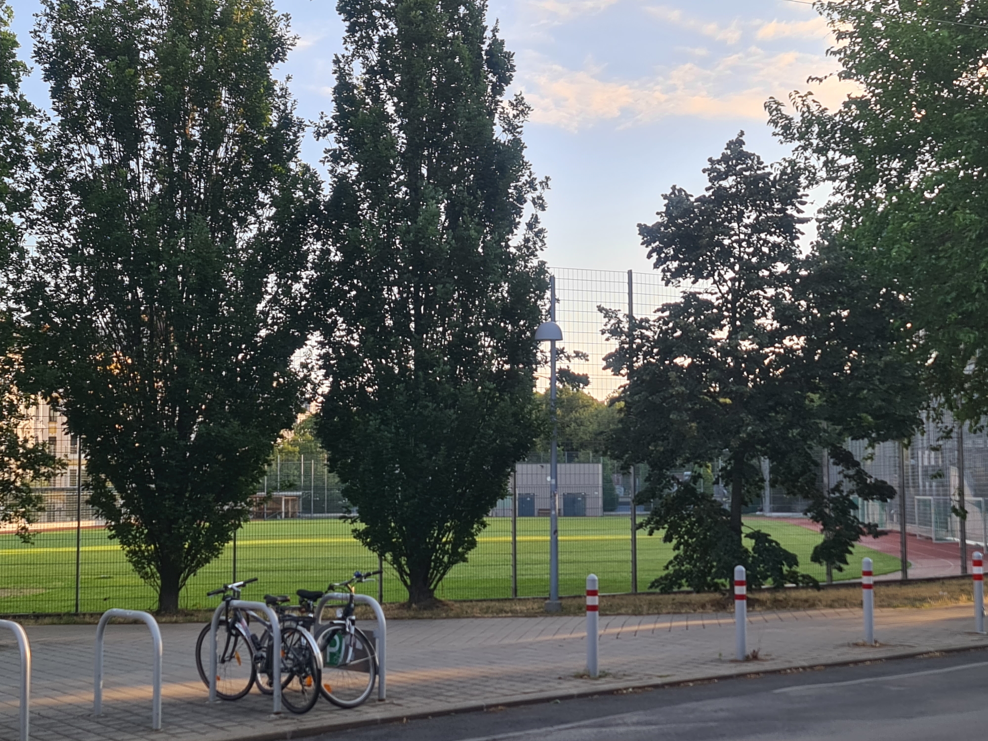 The image captures a scene on a paved area next to a fence surrounding a green field, likely a sports field. Several tall, leafy trees are prominent, with a clear sky above featuring some scattered clouds. Two bicycles are parked in a rack on the paved area in the foreground. The environment appears to be outdoors in a city. The presence of a sports field, trees, and a paved pathway suggests a park or recreational area within an urban setting. There are no people visible in the image. The lighting indicates it is either early morning or late afternoon. The overall impression is one of a peaceful, everyday scene in a city environment.